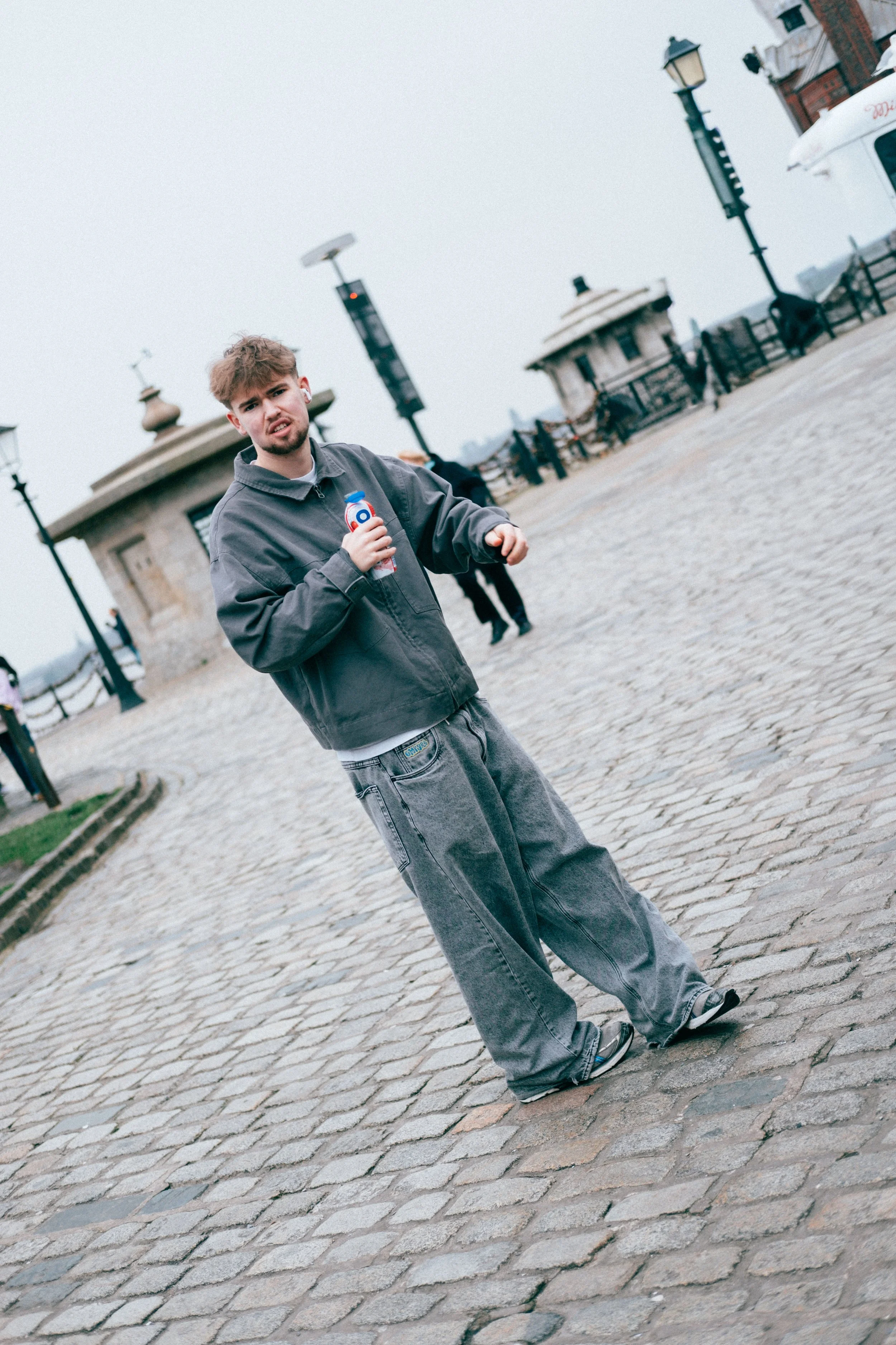 Young man with brown hair and beard standing on cobblestone street holding a water bottle, wearing gray pants, sneakers, and a dark jacket, with a cloudy sky and lampposts in the background.