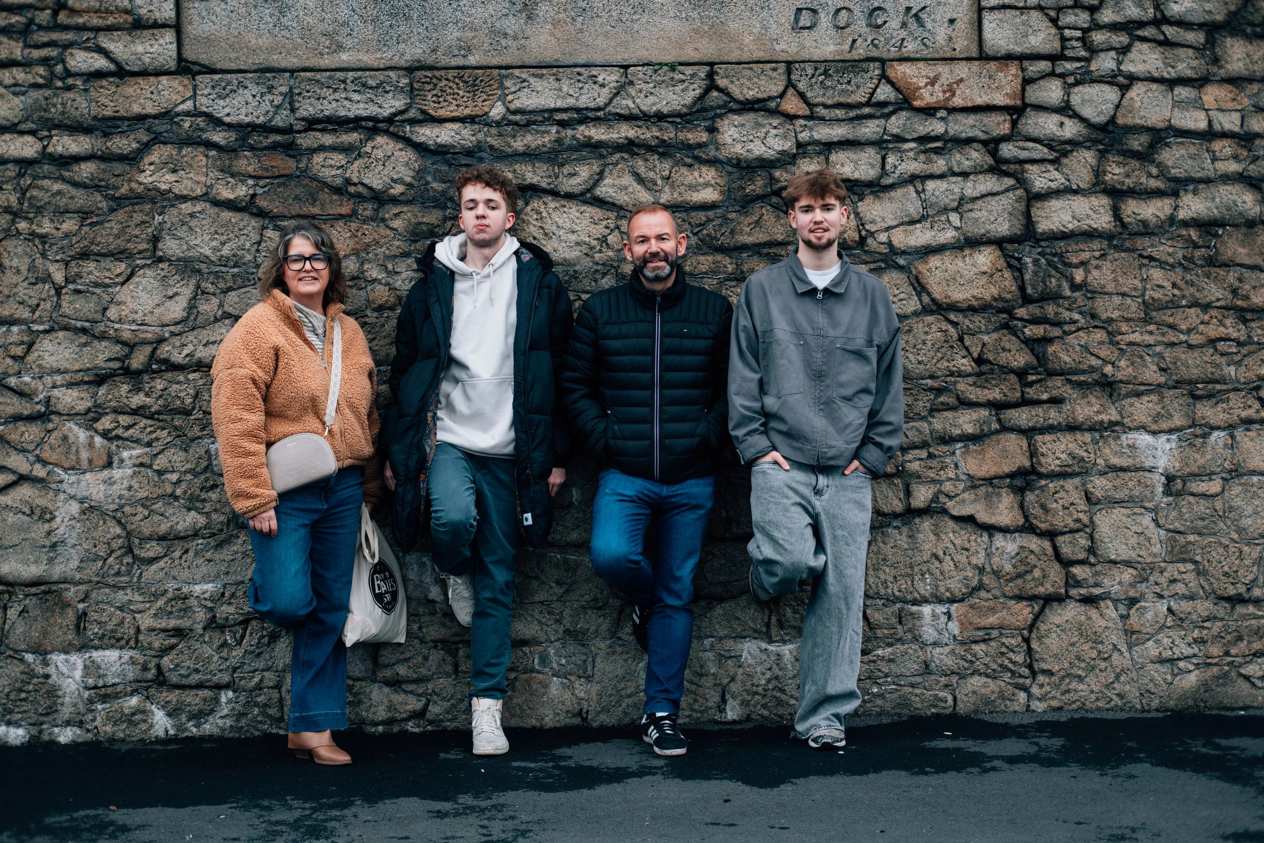 Four people standing in front of a stone wall, with one woman and three young men, all dressed in casual jackets and pants, smiling and posing for the photo.