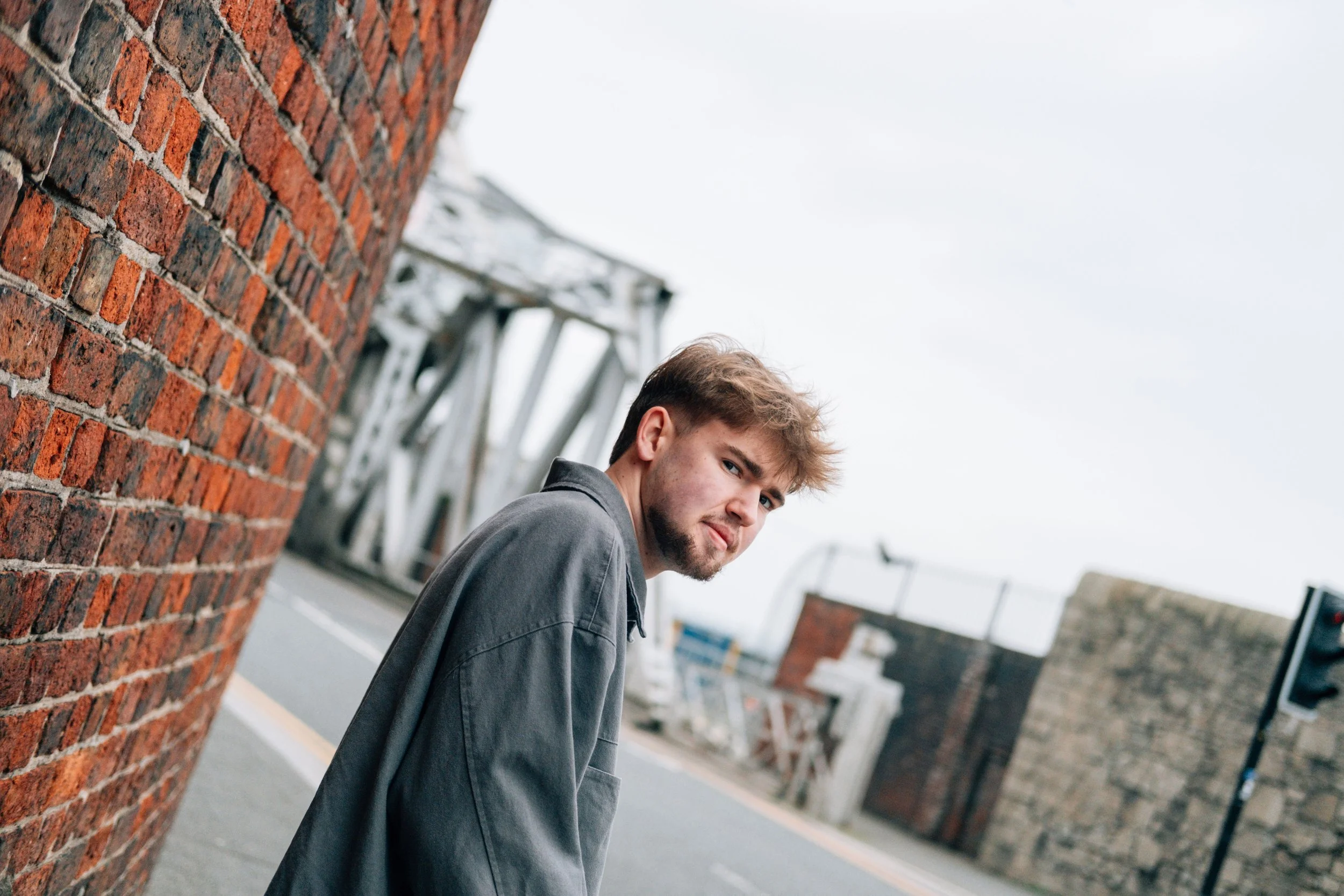 Young man with light brown hair and beard standing outdoors next to a red brick wall, looking over his shoulder.