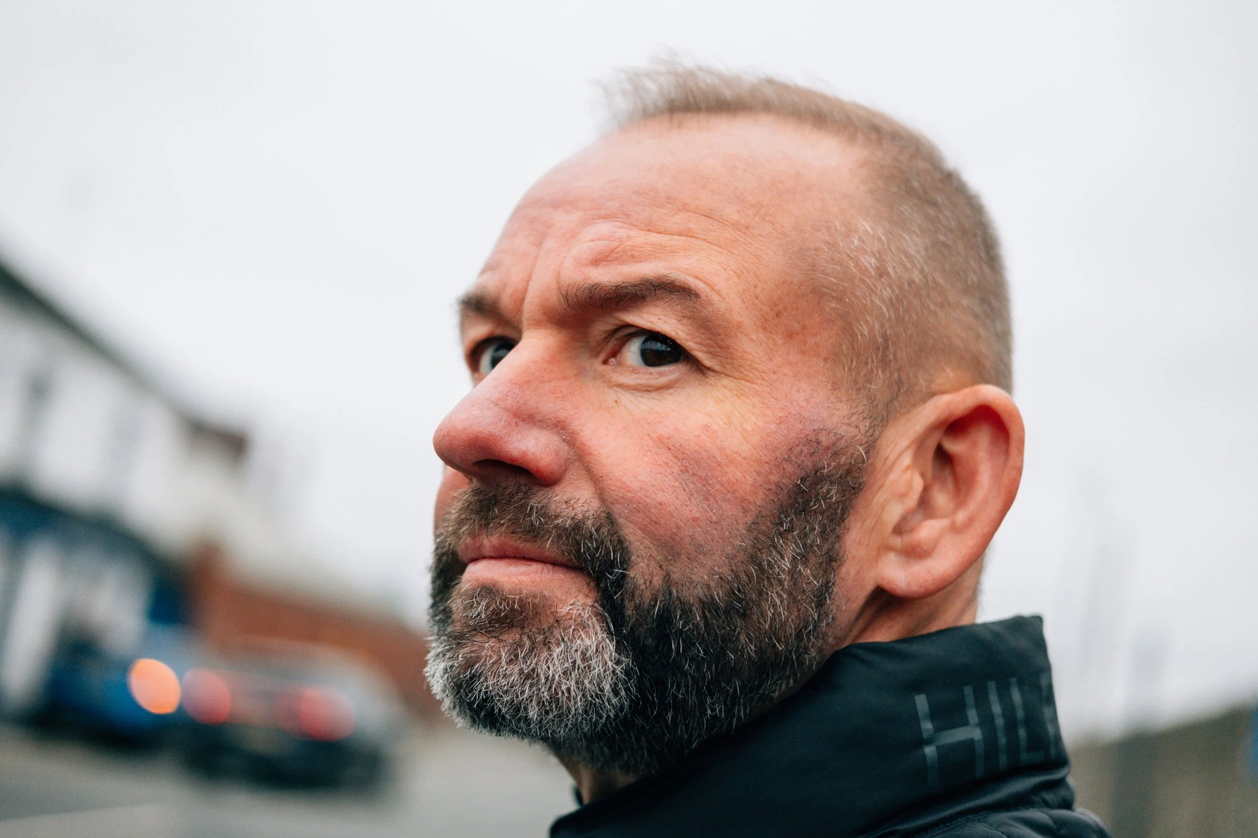 Close-up of a middle-aged man with a beard and short hair, wearing a black jacket, standing outdoors on a cloudy day with blurred buildings in the background.