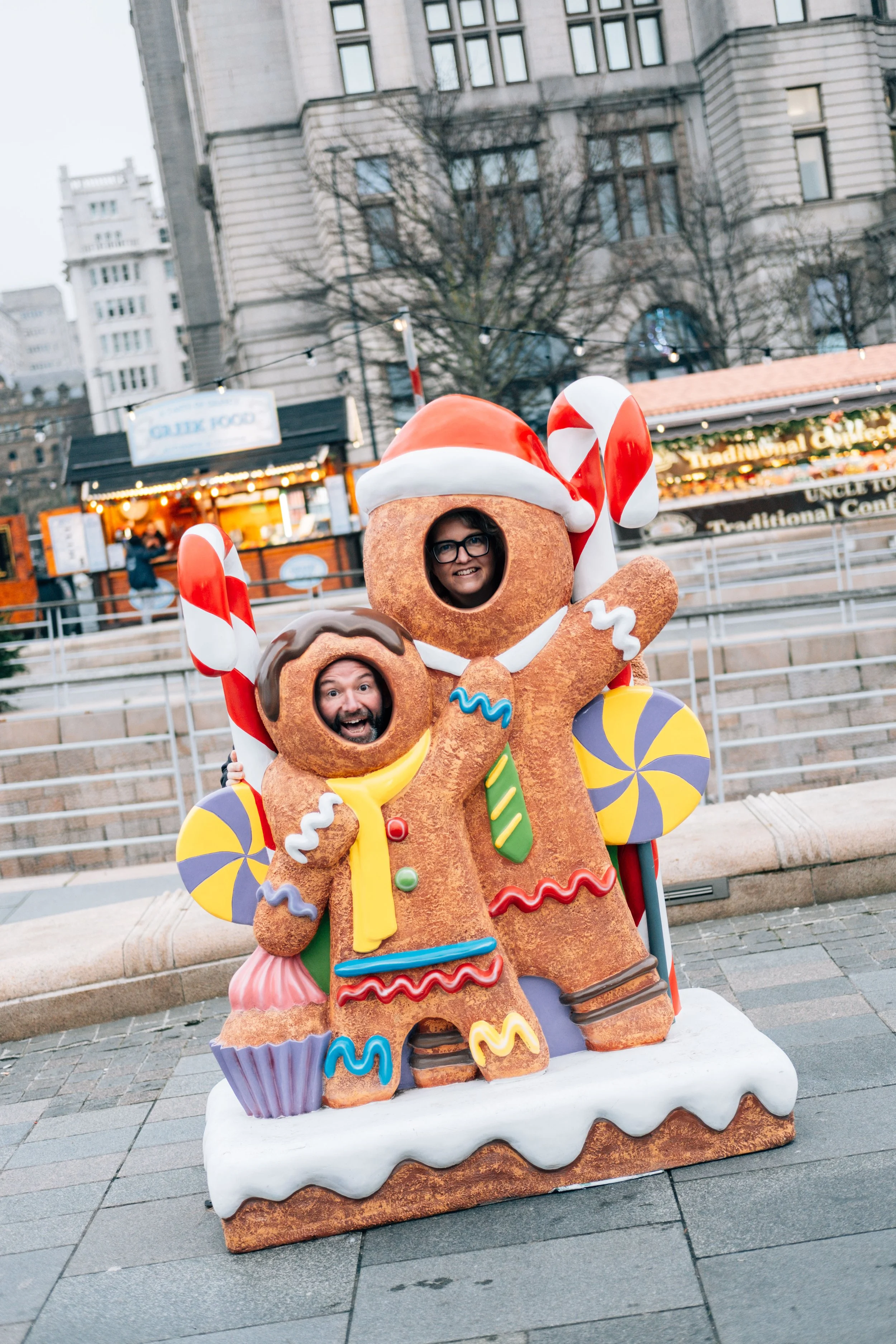 Two people with faces seen through cookies on a holiday gingerbread stand, decorated with Christmas-themed accessories at an outdoor event.