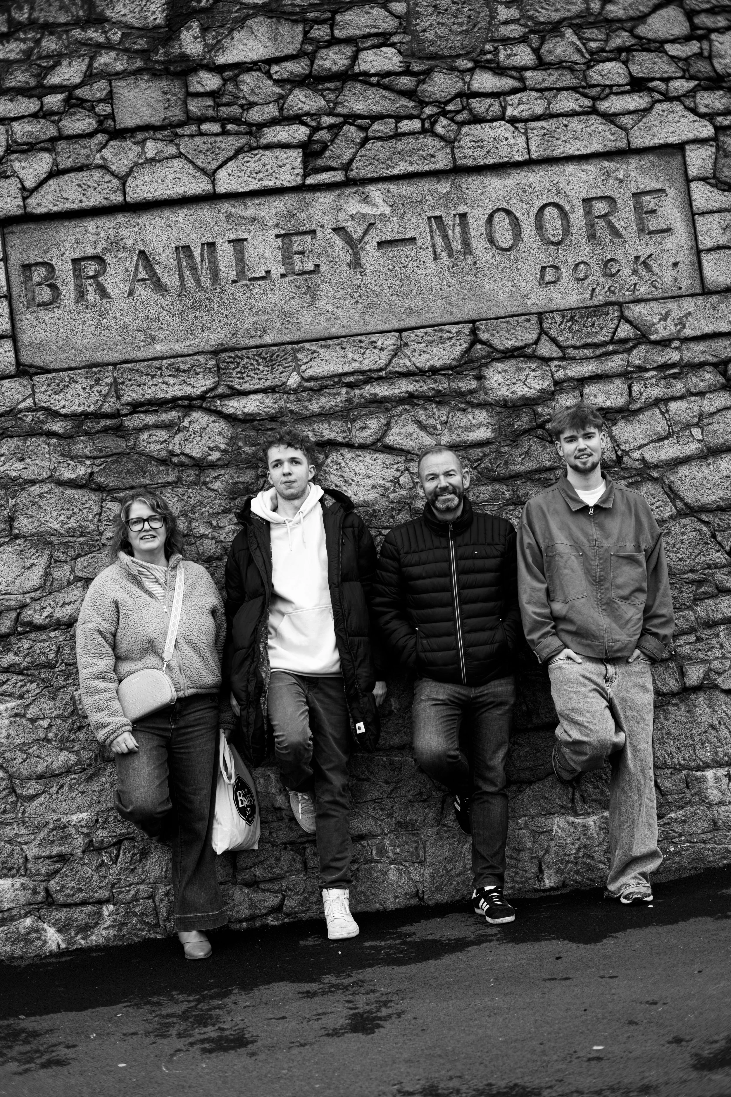 Group of four people standing against a stone wall with a large engraved sign that reads "Bramley-Moore Dock 1825."
