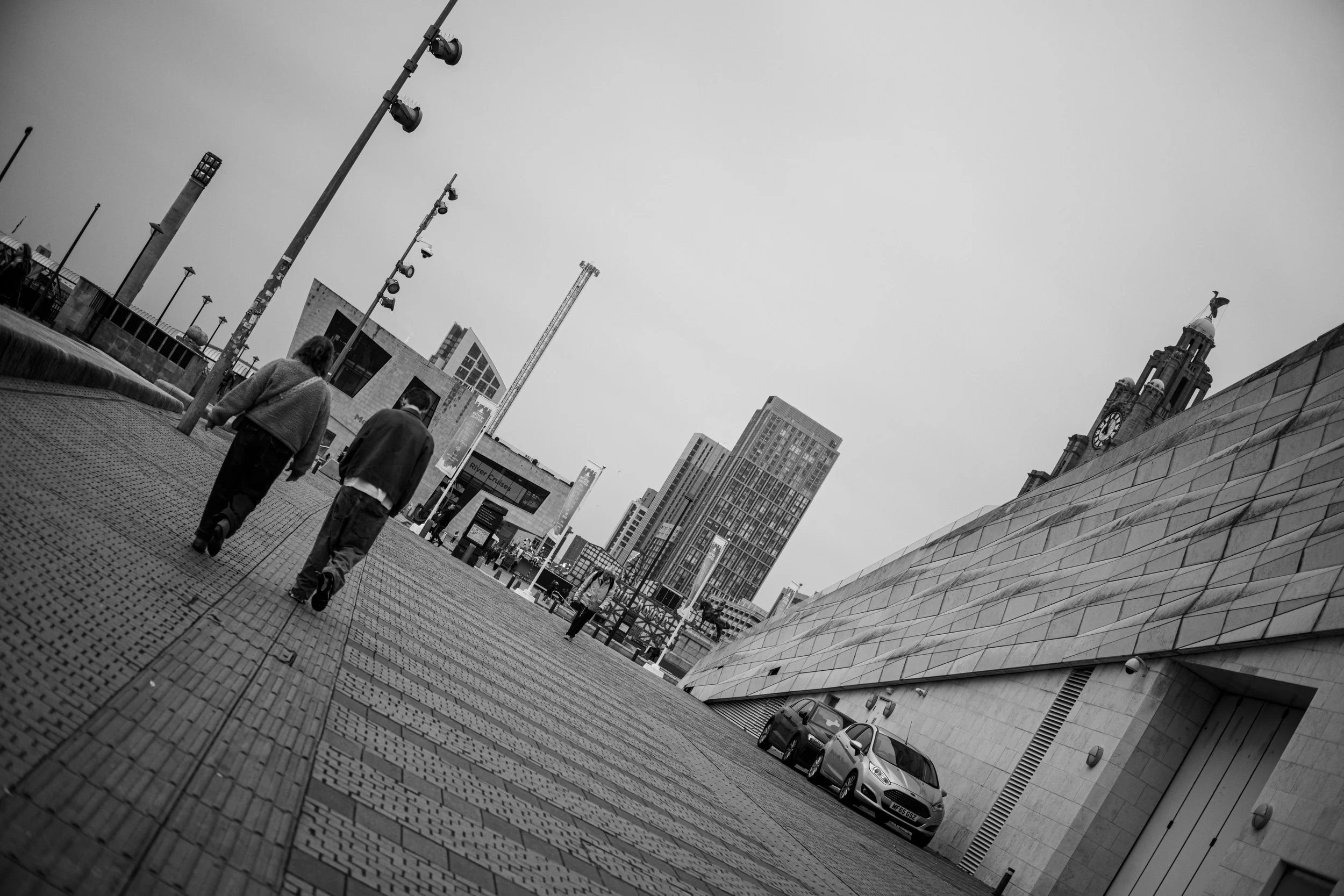 Black and white photo of an urban street scene with two people walking, modern buildings, a partially tilted perspective, cars parked along the sidewalk, and a clock tower in the background.