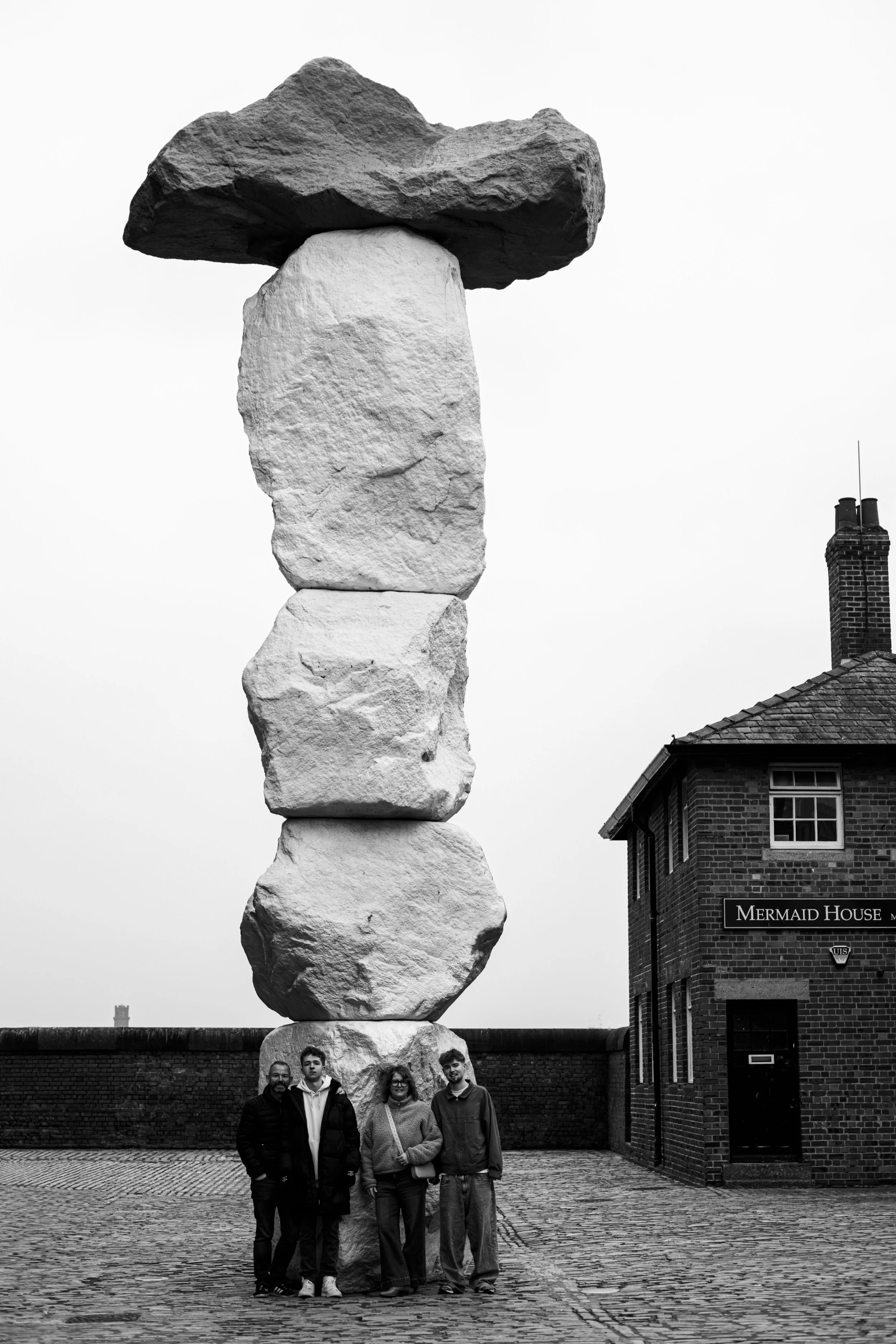 Four people standing under a large stone sculpture resembling a balanced stone formation, with a building labeled 'Mermaid House' nearby.