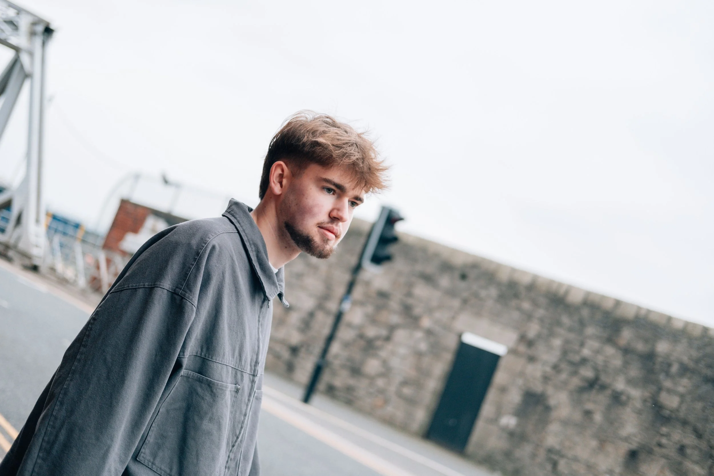 Young man with light brown hair and beard wearing a gray jacket standing on a city street near a brick wall and traffic light.