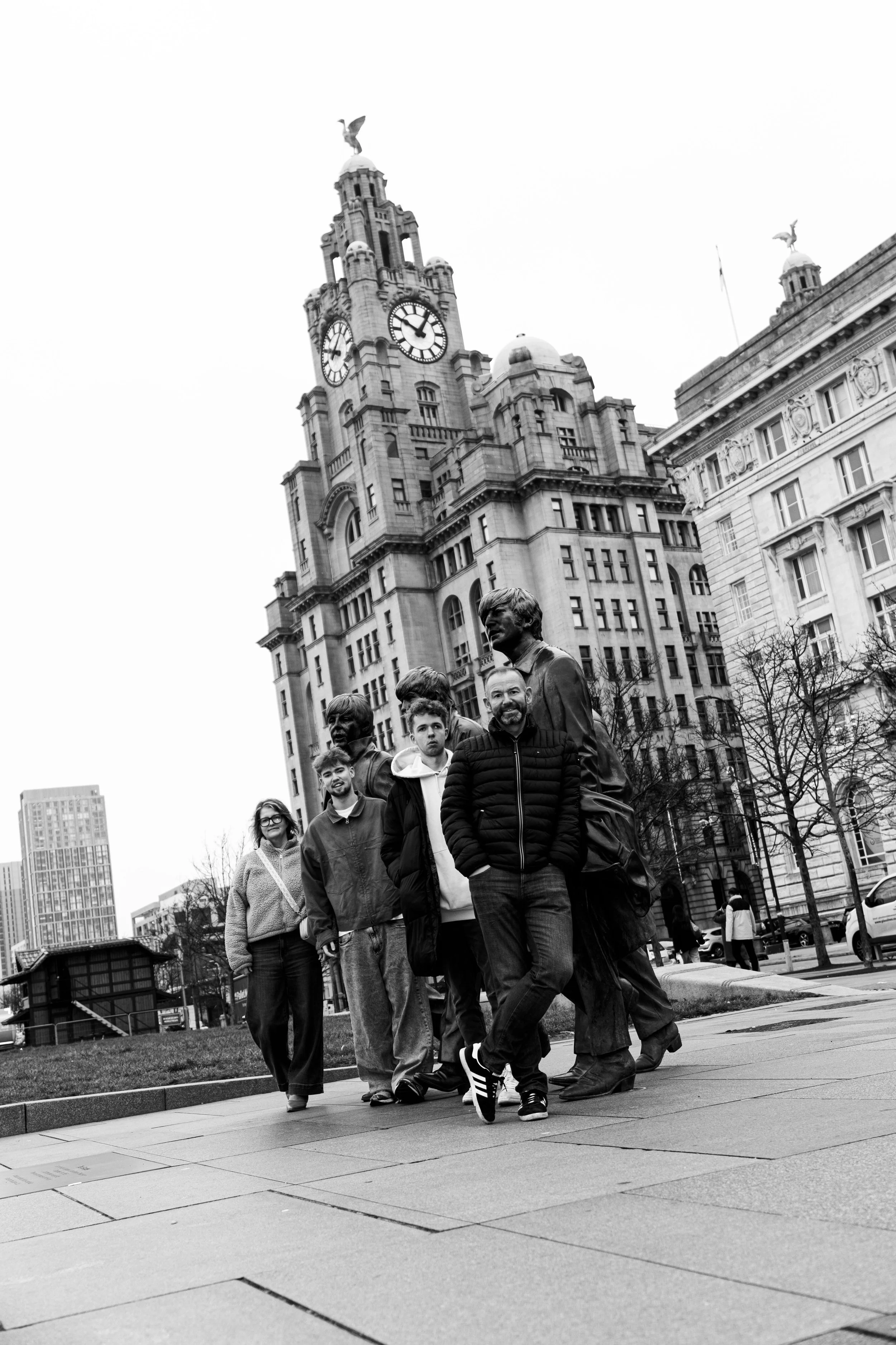 A group of people walking past statues of historical figures in front of a large clock tower in an urban setting