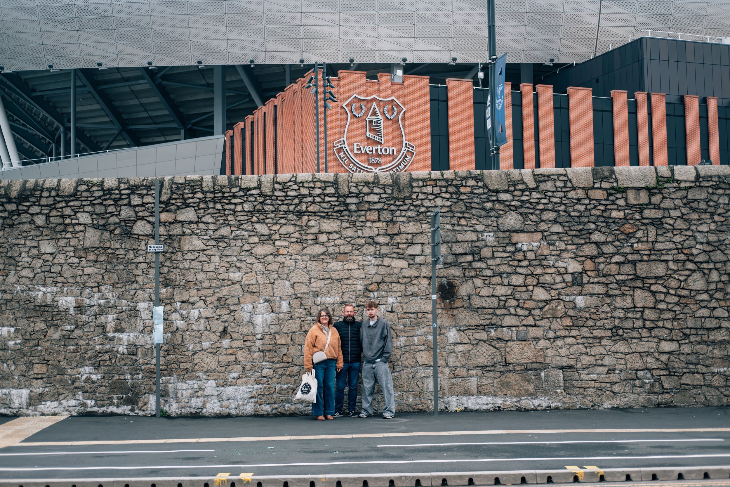 Three people standing in front of a stone wall outside a stadium with an Everton Football Club crest and a modern building behind them.