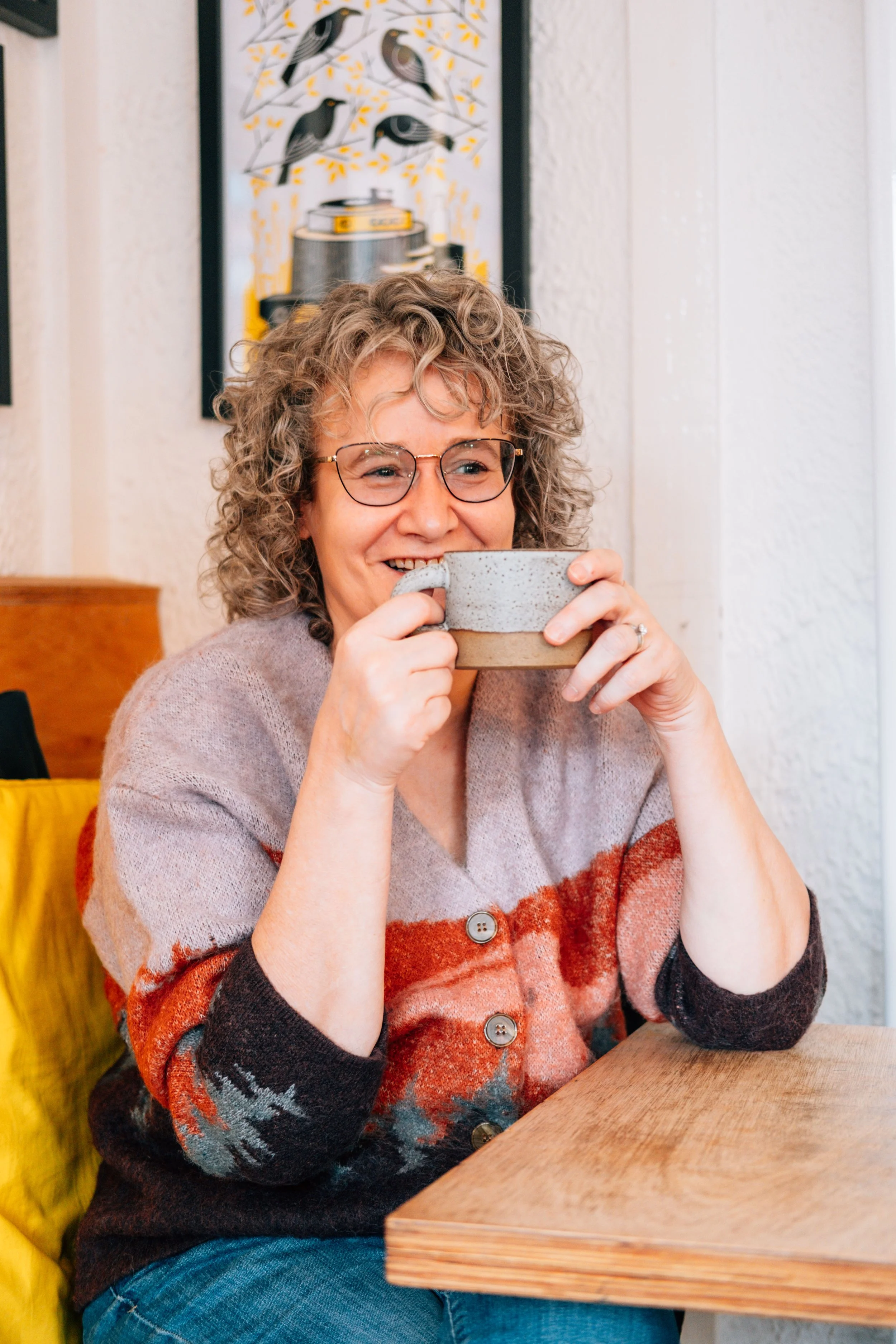 A woman with curly hair and glasses, smiling and holding a mug, sitting at a wooden table in a cozy room.