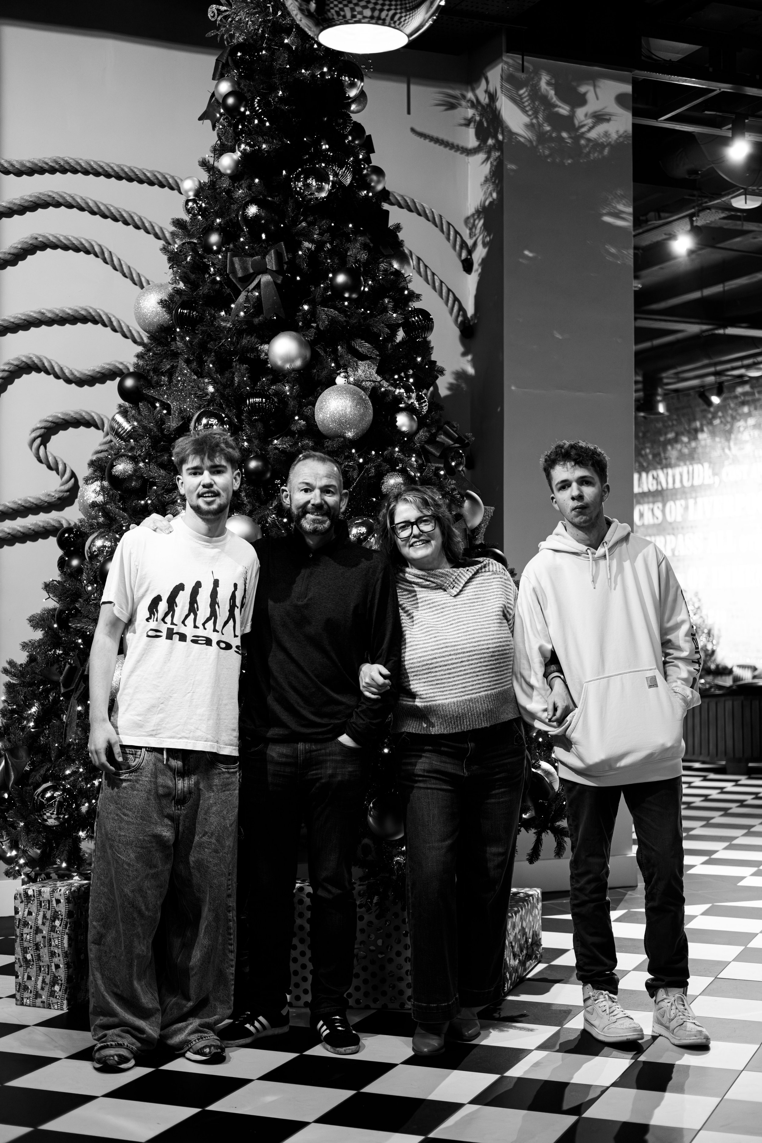 A family of four standing in front of a decorated Christmas tree, with presents underneath, in an indoor setting.