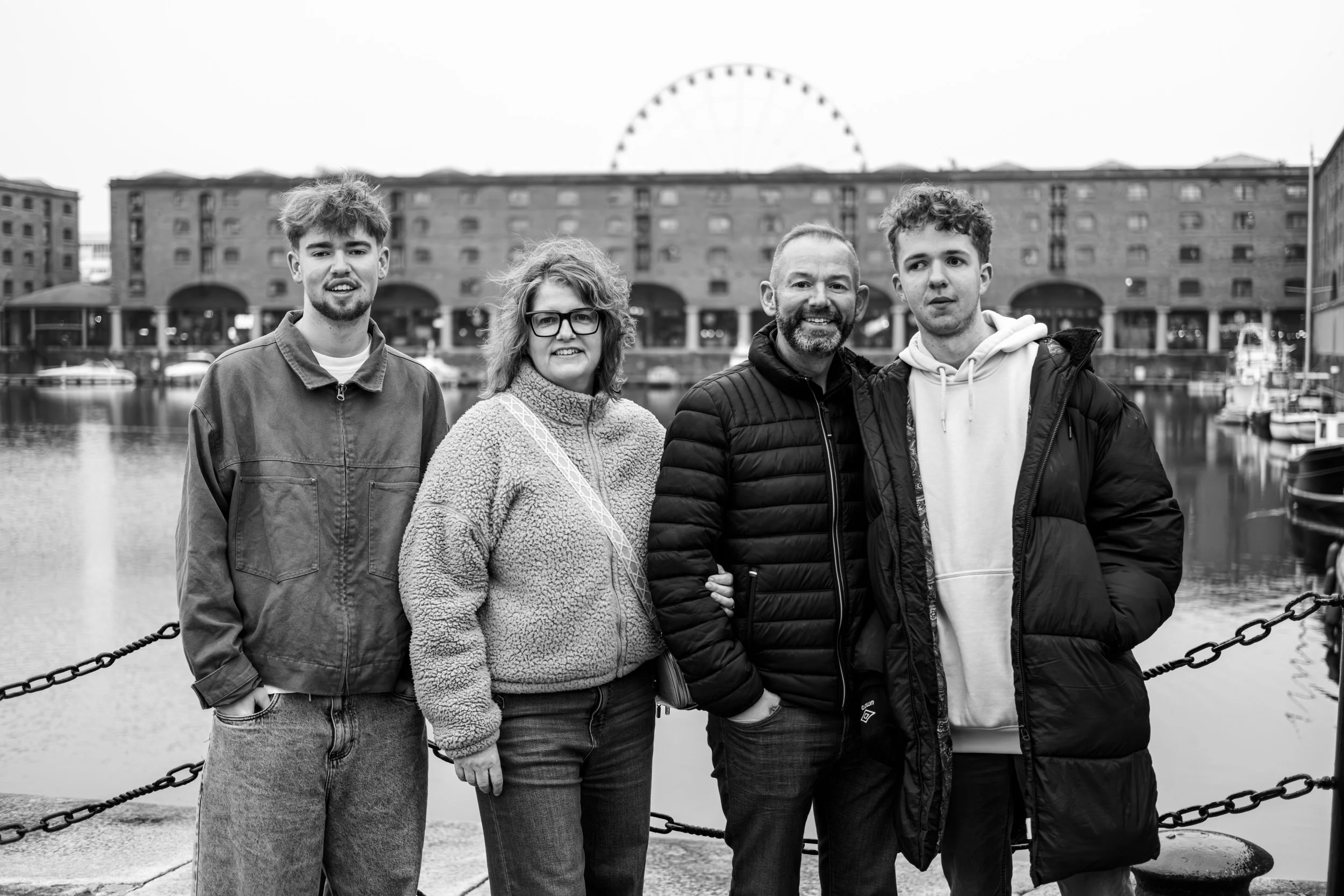 A black and white photo of a family of four standing by a harbor with boats and a cityscape in the background, including a large building and a Ferris wheel.