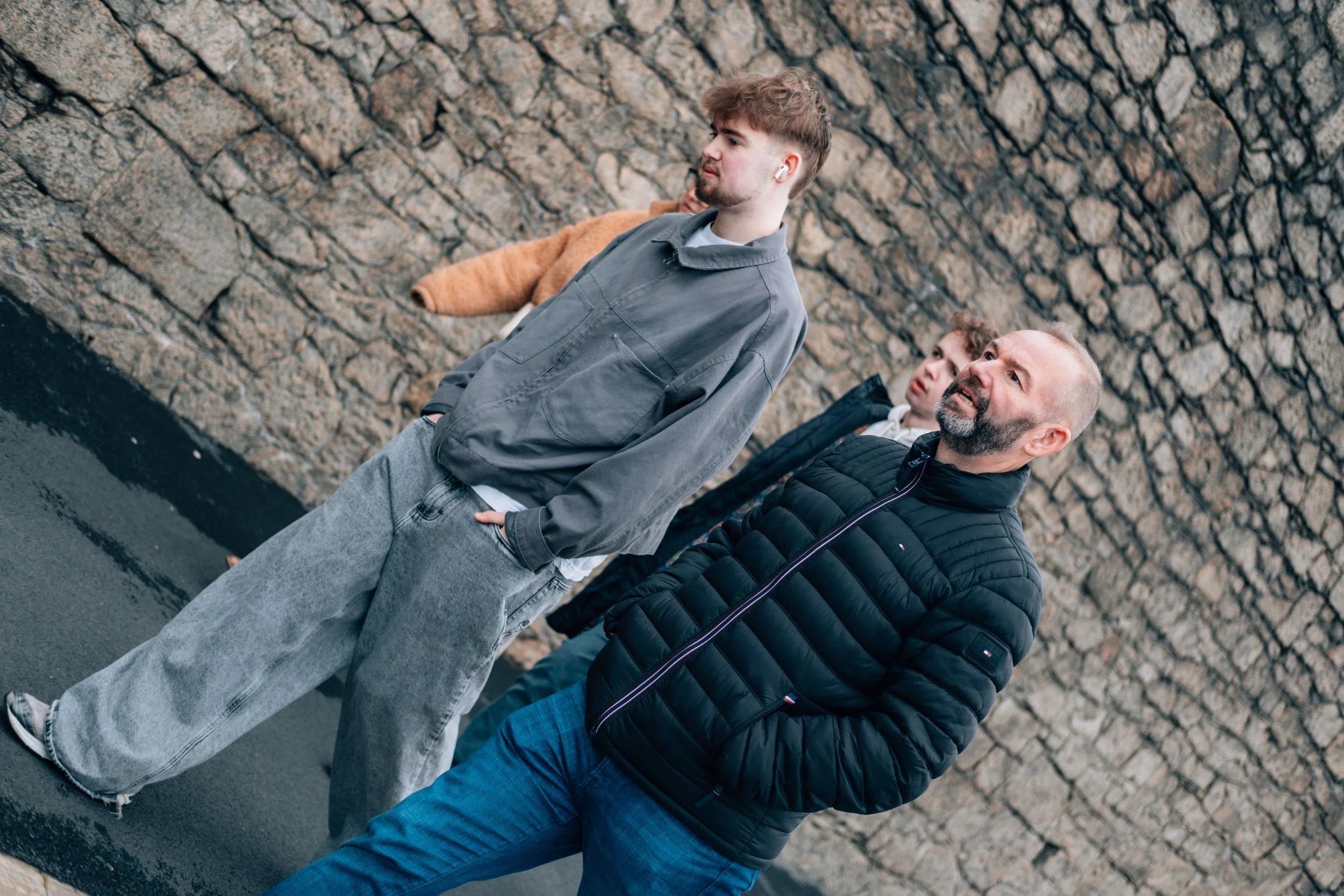 Four people walking in front of a stone wall, including a man with a beard wearing a black puffer jacket and two young men, one with a gray jacket and one with a brown coat, walking together outdoors.