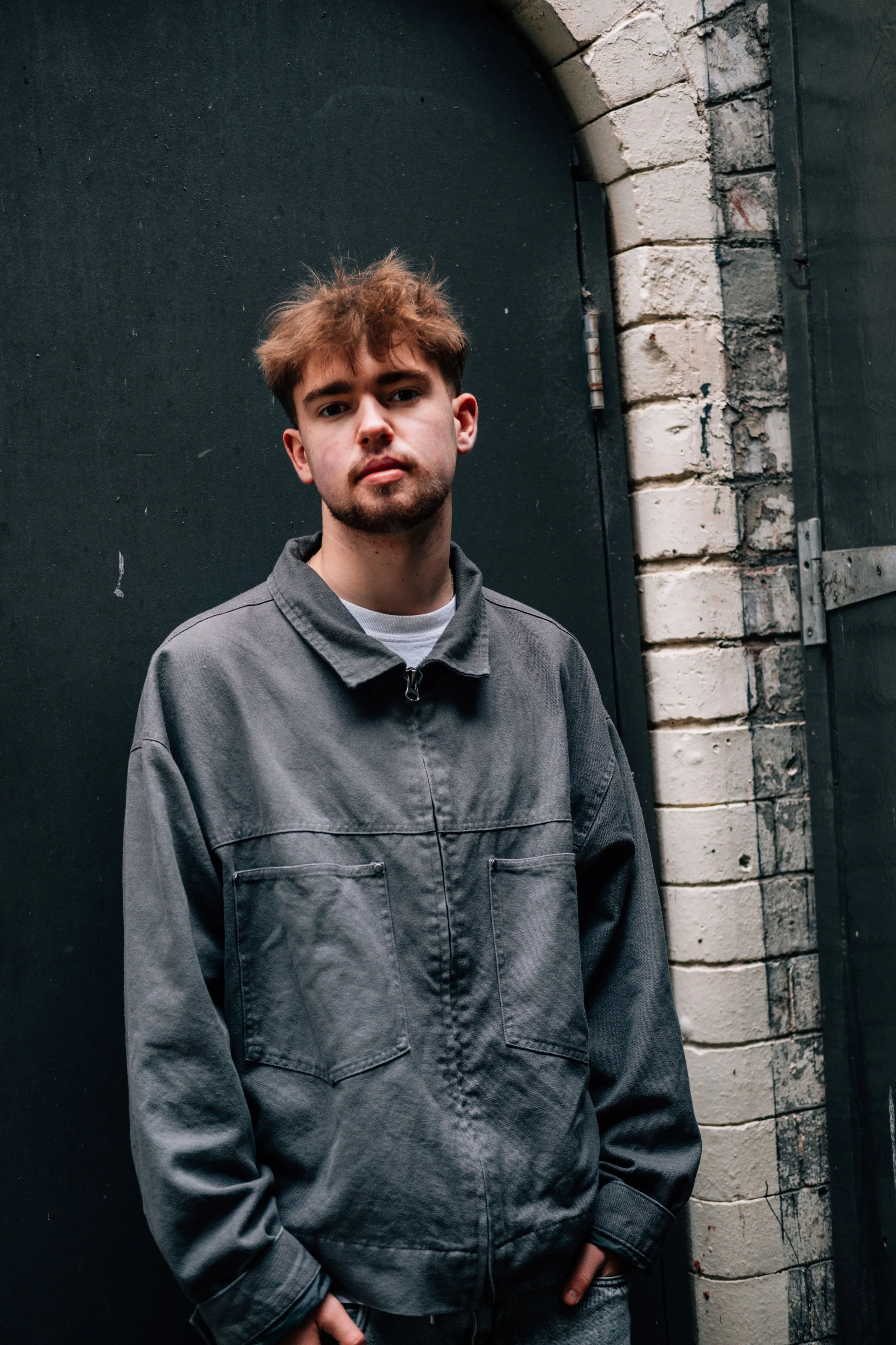 A young man with brown hair and a beard standing in front of a dark door and brick wall, wearing a gray jacket and looking at the camera.