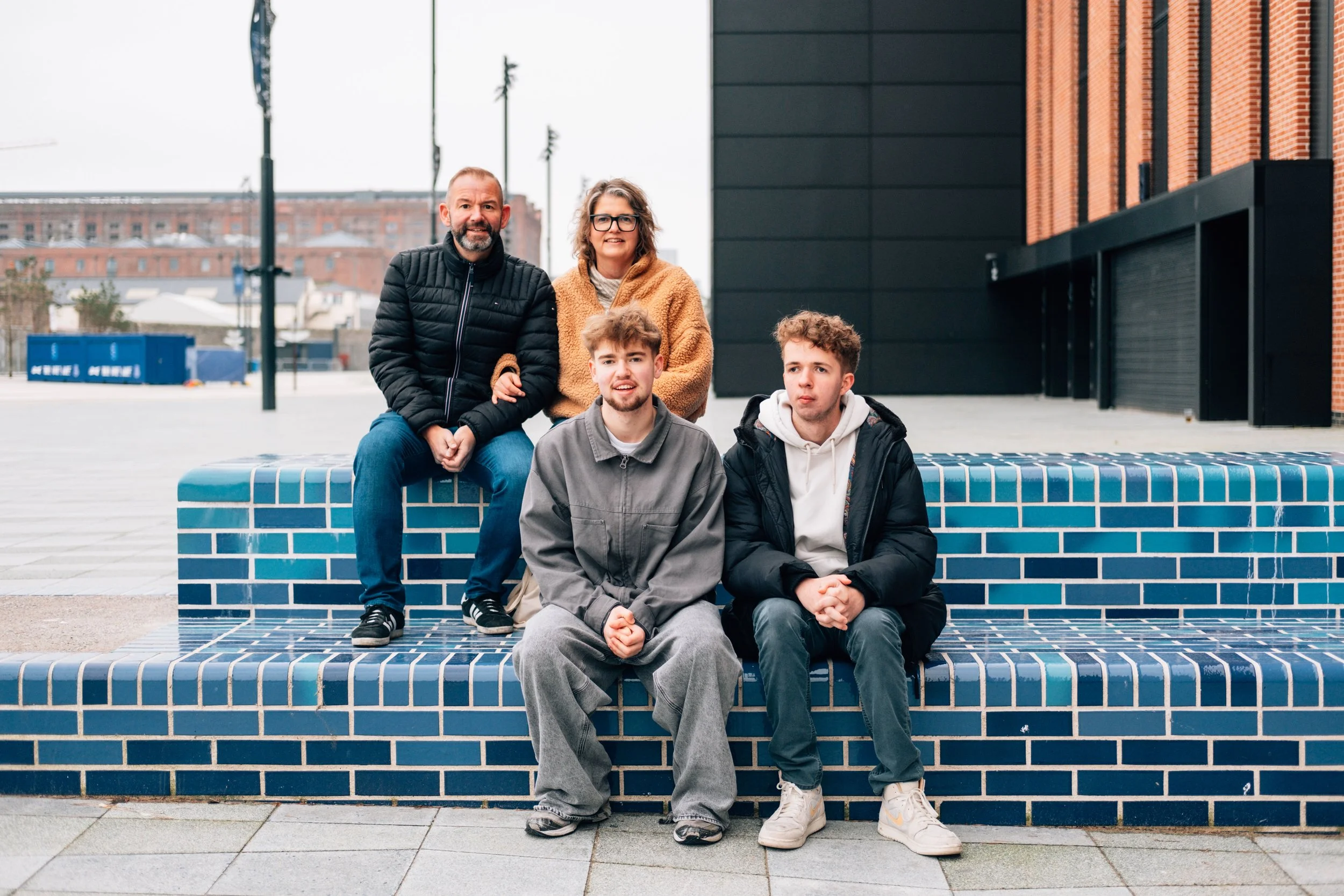 Group of five people sitting on a blue tiled bench outdoors in an urban area with modern buildings, dressed in casual jackets and hoodies.