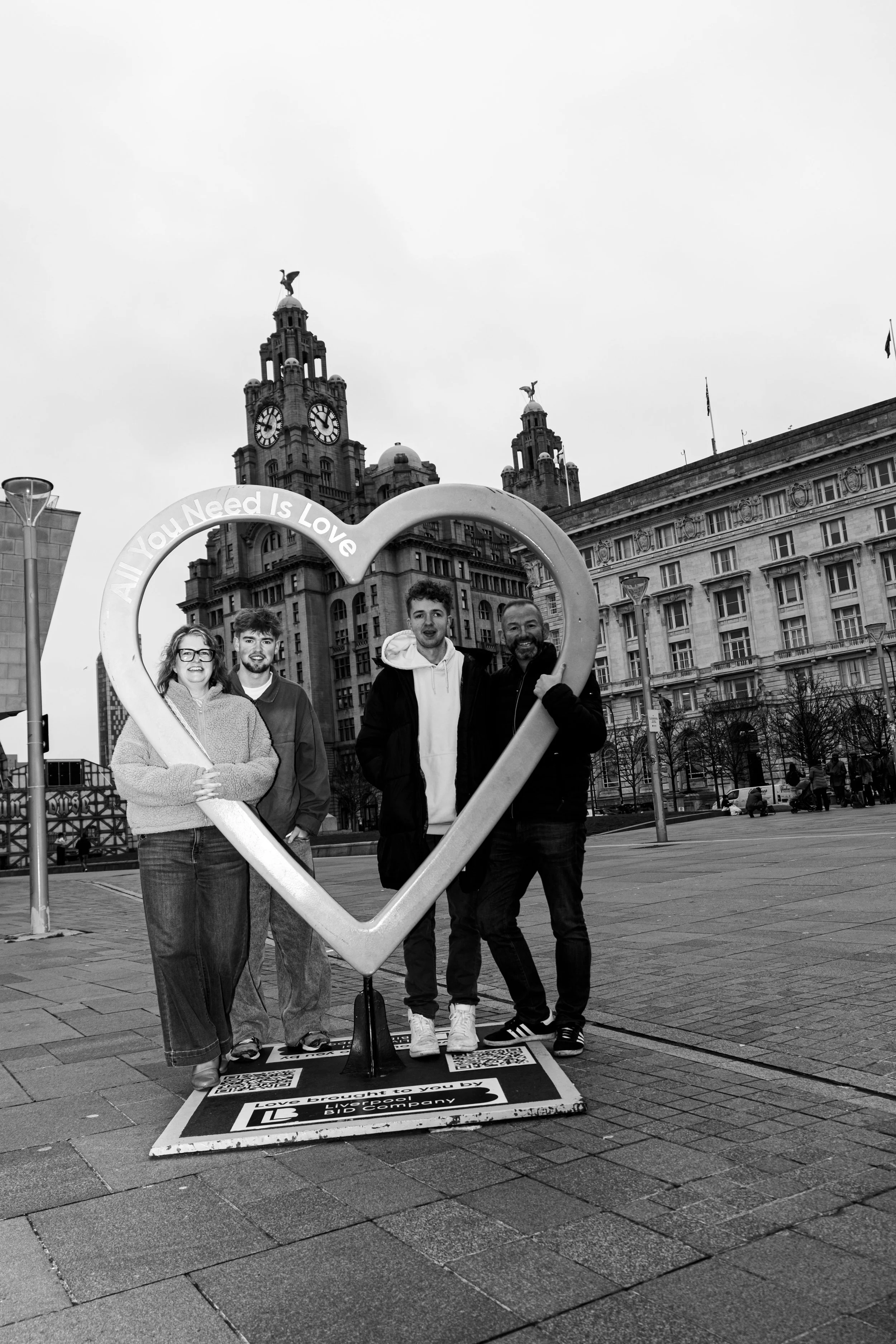 Four people standing behind a large heart-shaped frame with the words "All You Need Is Love" written on top, in an urban square with historic buildings and clock towers in the background.