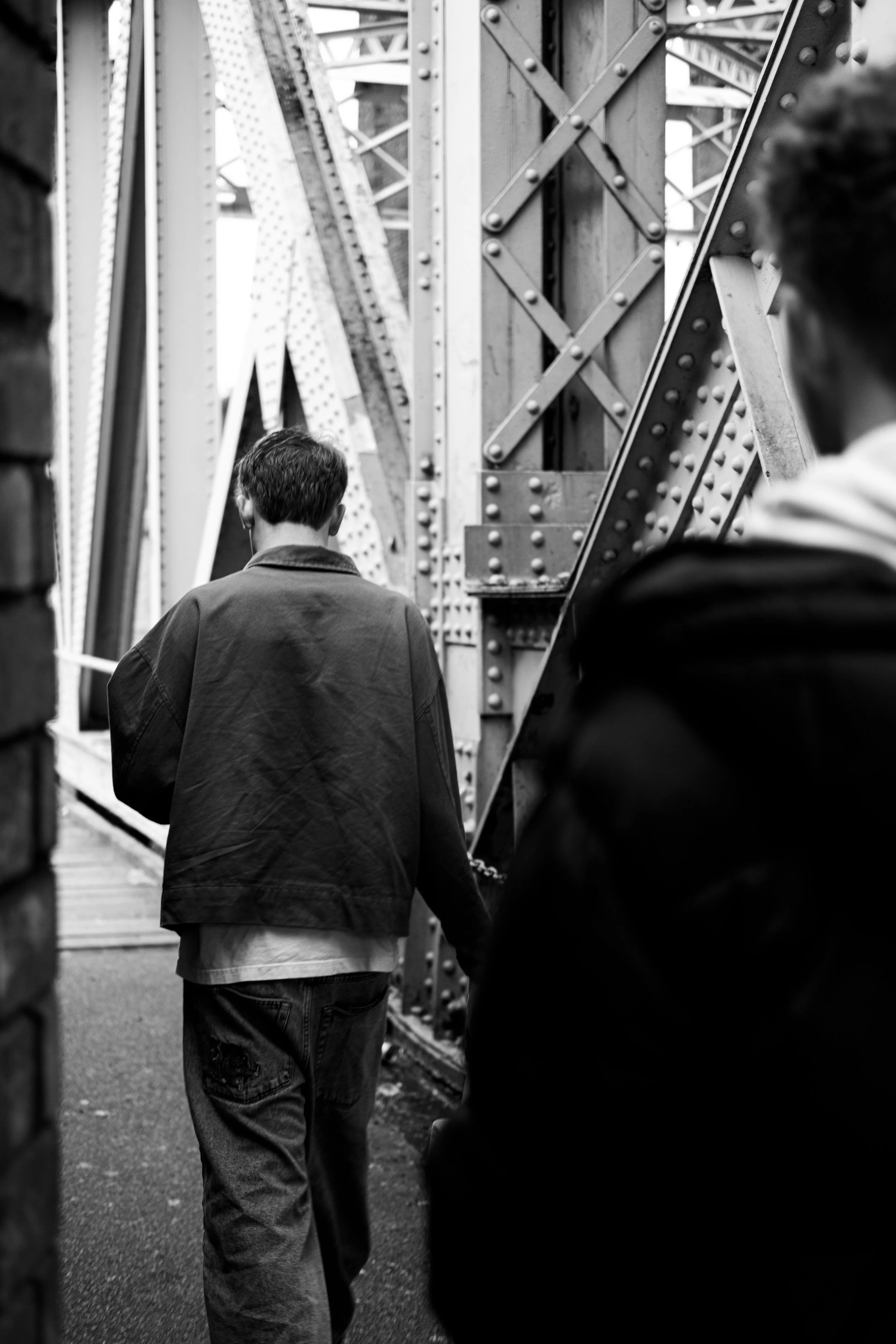 Two men walking through a steel bridge, one is seen from behind wearing a jacket and jeans, and the other is partially in view on the right, wearing a jacket with a hood.