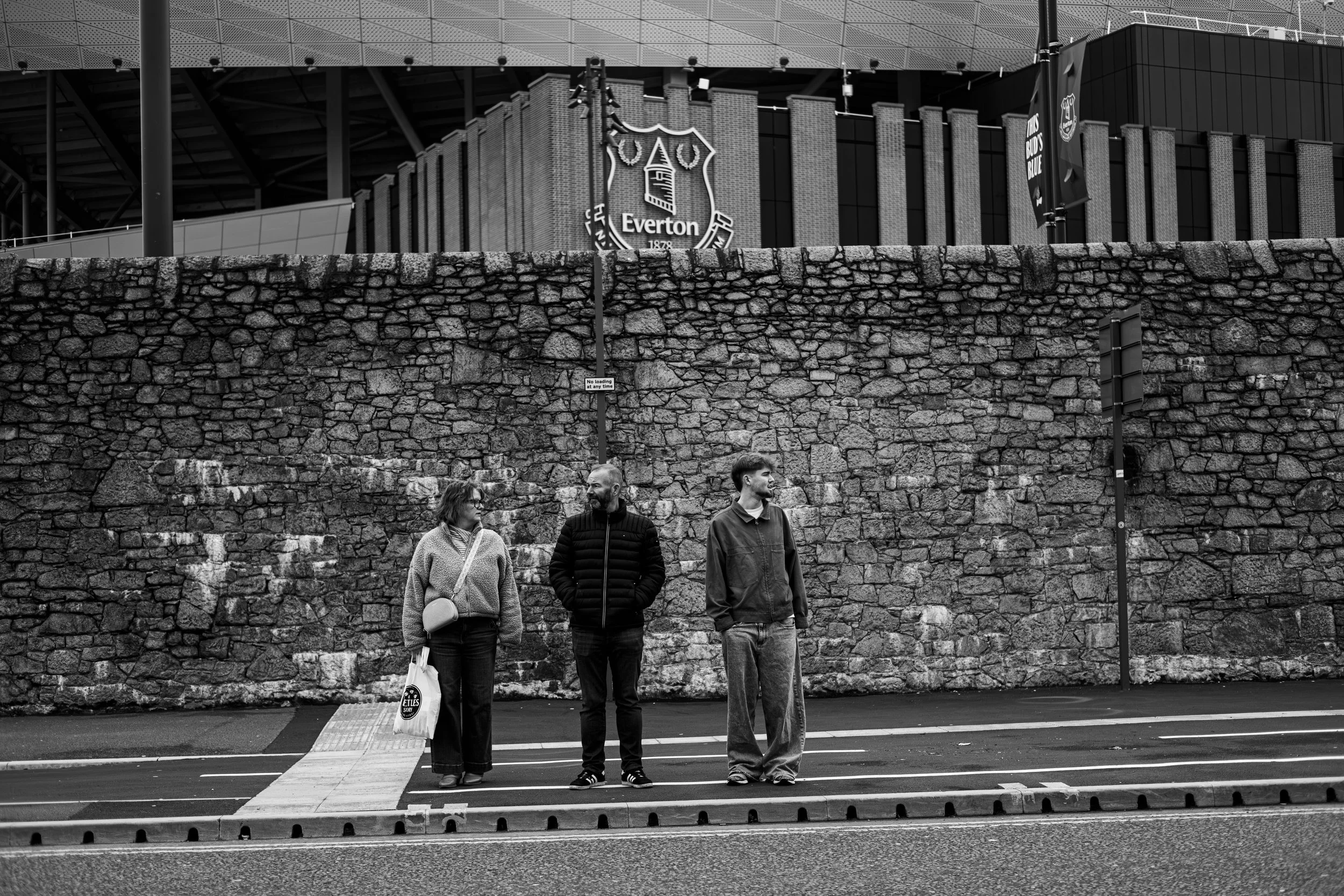 Three people standing on crosswalk in front of brick wall, with Everton football stadium in background