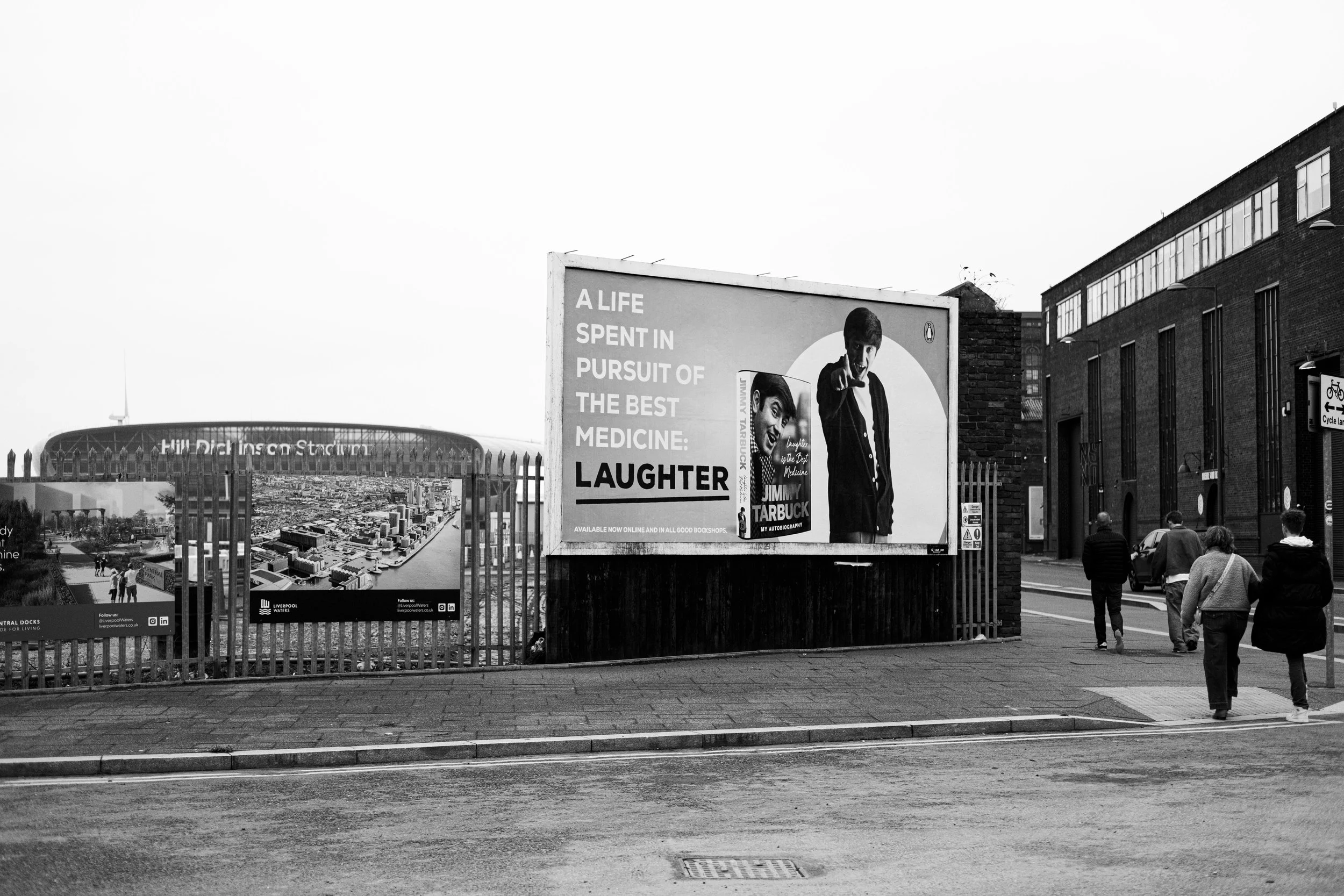 Black and white street scene with a billboard featuring two men, one pointing at the camera. Sign reads, 'A life spent in pursuit of the best medicine: laughter'. Three pedestrians walk along the sidewalk, and a brick building is visible on the right