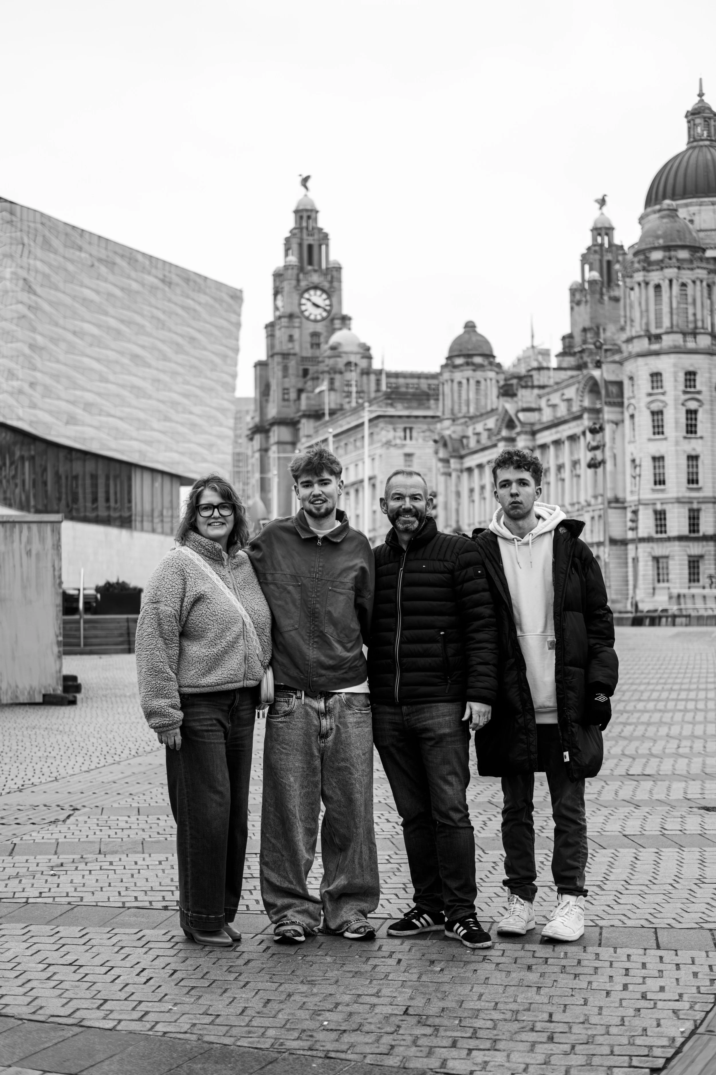 Four people standing together in a city square with historic buildings, including a clock tower and domed structures, in the background.
