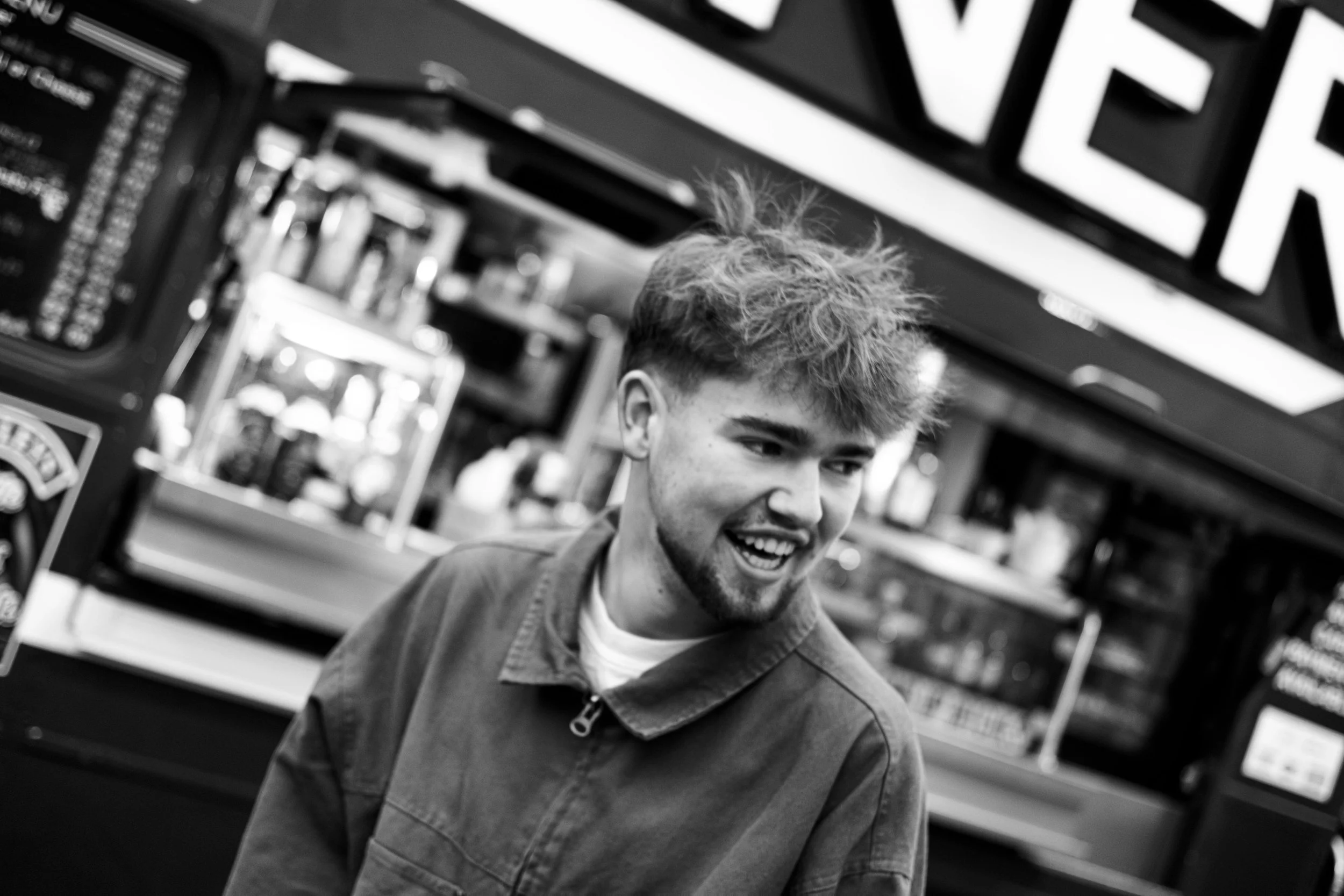 A young man with messy hair and a beard smiling, standing outside at a convenience store with vending machines in the background, in black and white.