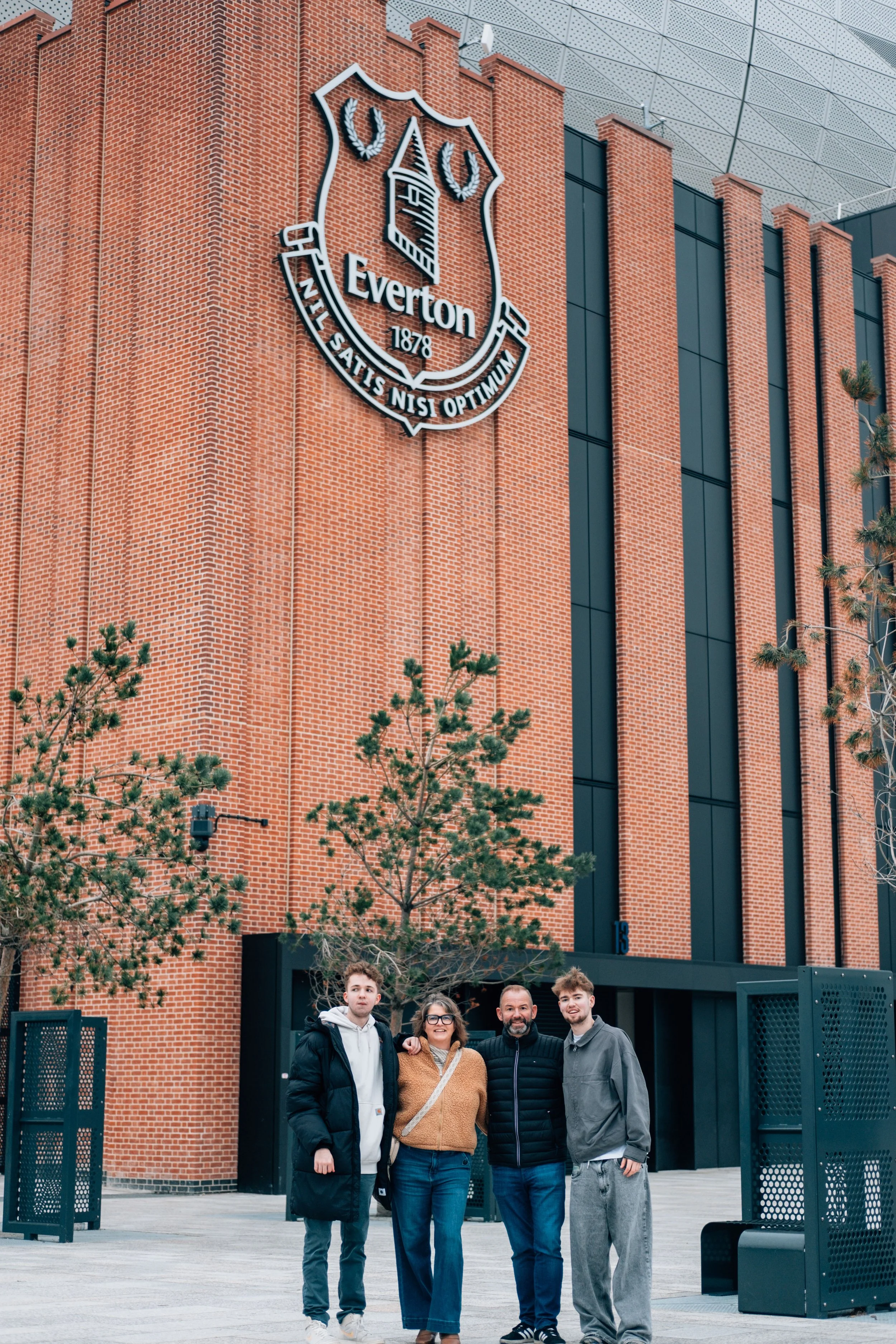 Four people standing in front of a red brick building with the Everton Football Club crest on it.