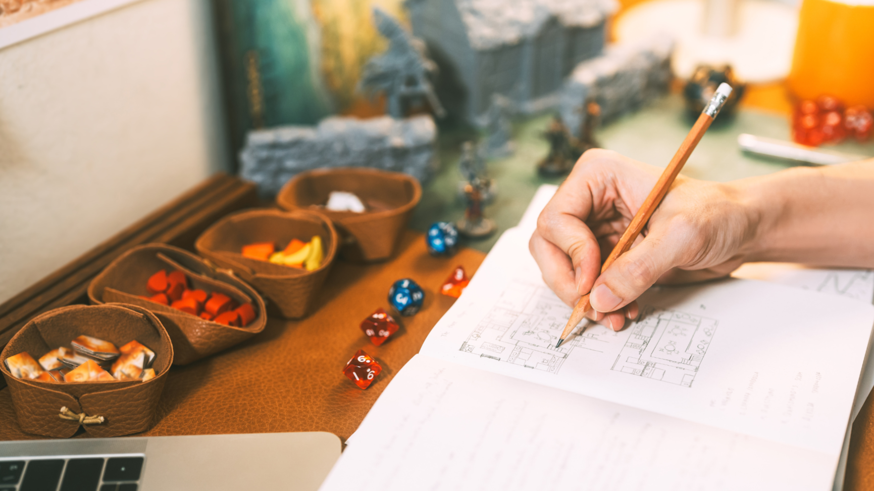 A person is drawing or writing on blueprints or floor plans with a pencil at a desk. There are several small bowls with colorful tokens or game pieces, some scattered on the desk. In the background, there are miniature figurines and rocks, possibly for tabletop gaming.
