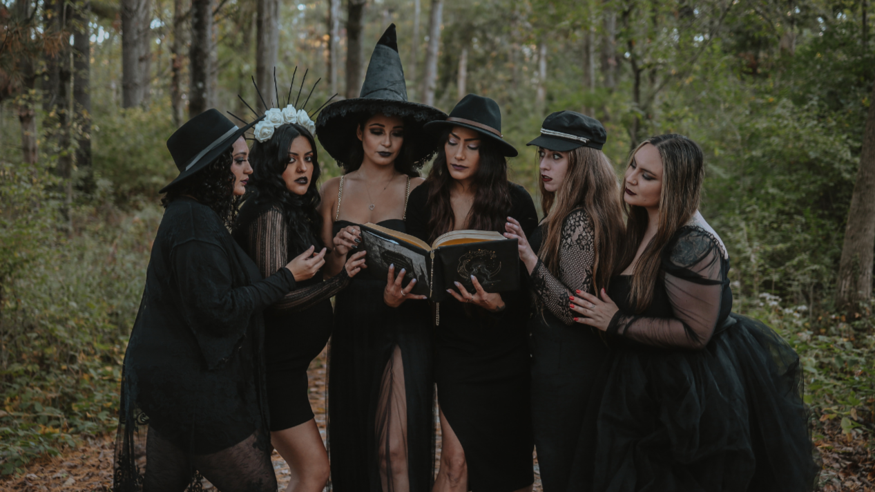 Six women in black witch costumes and hats gather in a forest, reading a spellbook together.