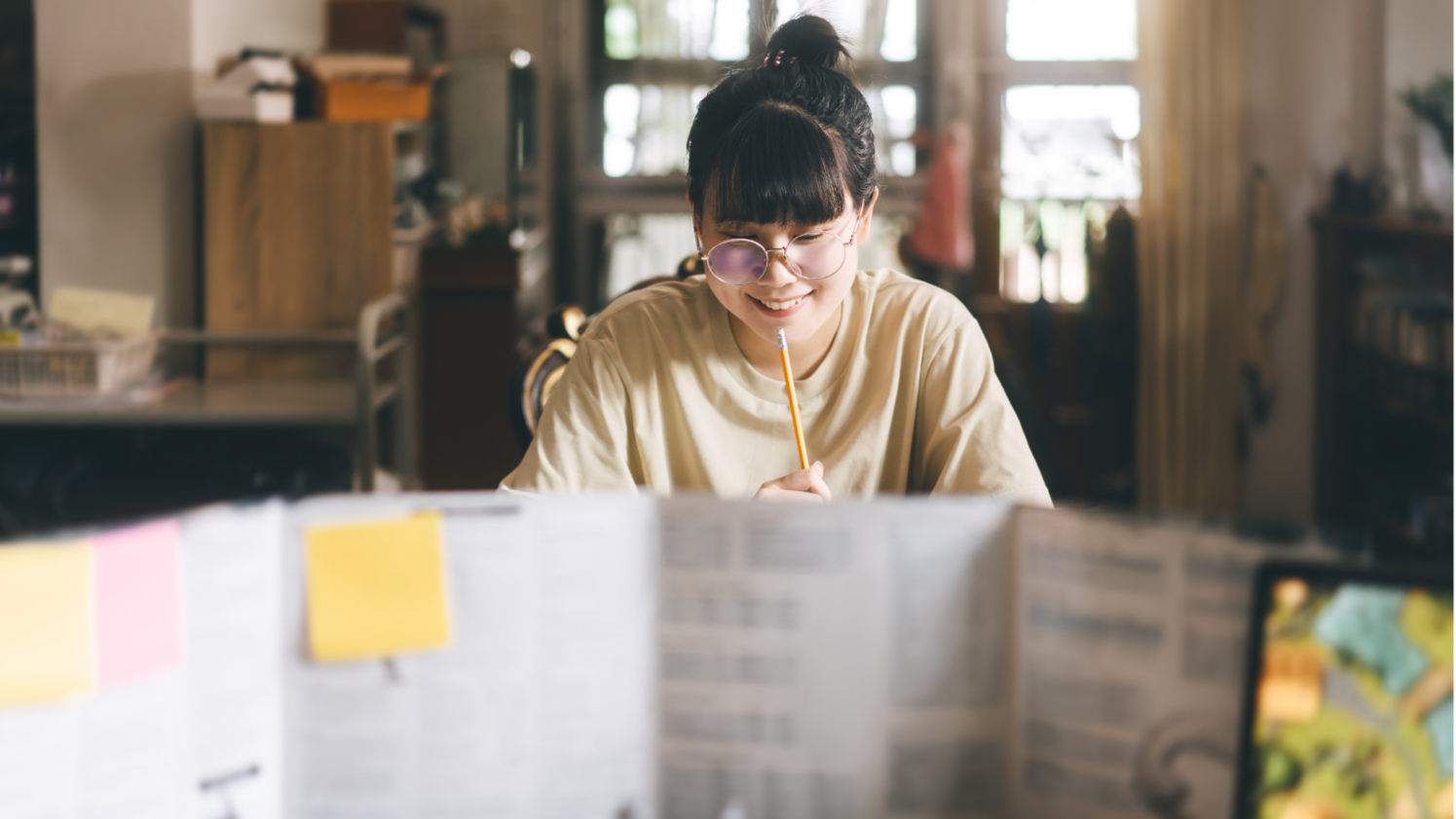 A smiling young woman with glasses and a bun hairstyle, sitting at a desk with open books and notes, holding a pencil near her mouth, in a well-lit room with large windows.