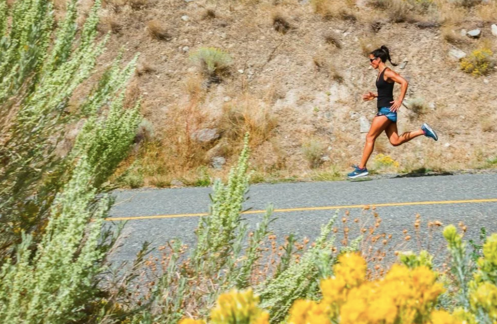 A woman jogging on a paved road in a hilly, dry landscape with yellow flowers in the foreground.