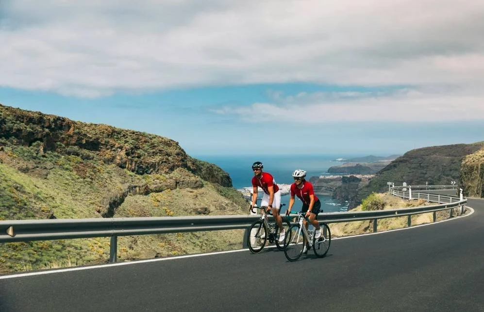 Zwei Radfahrer, die gemeinsam auf einer Küstenbergstraße fahren, symbolisieren die intrinsische und extrinsische Motivation im Ausdauertraining.
