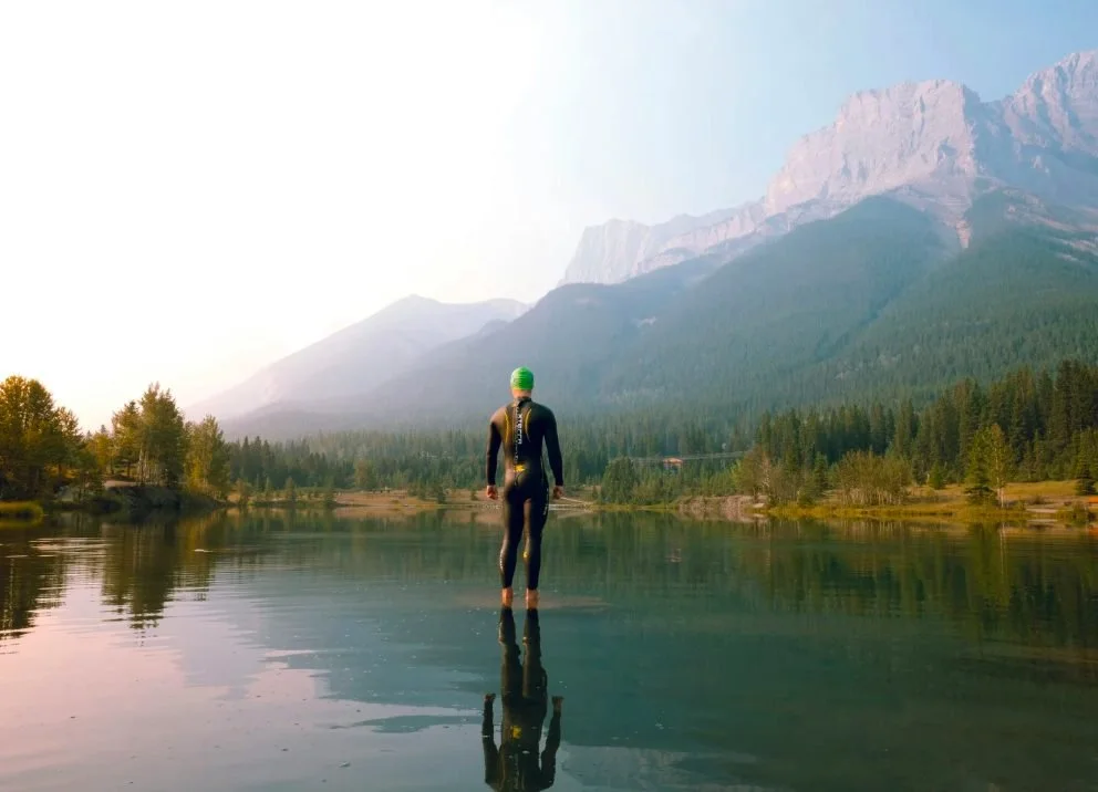 Athlete standing in still water facing mountains, representing self sabotage and the discomfort of moving forward in endurance training.