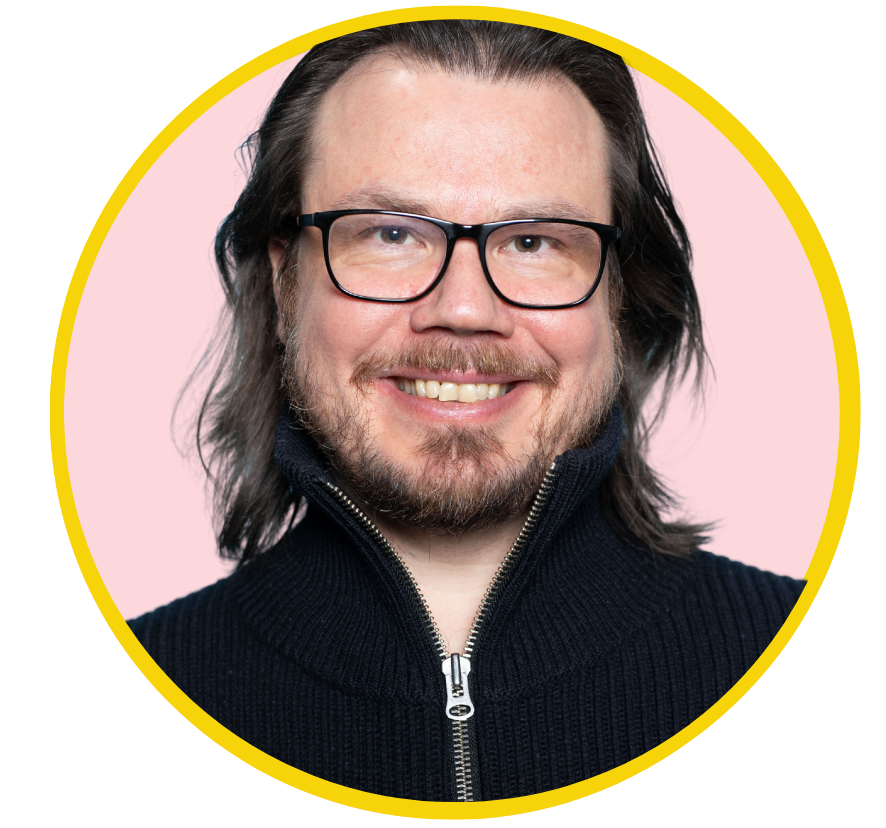 Smiling man with long dark hair, glasses, beard, wearing a black zip-up sweater, pink background. Lauri Järvilehto.
