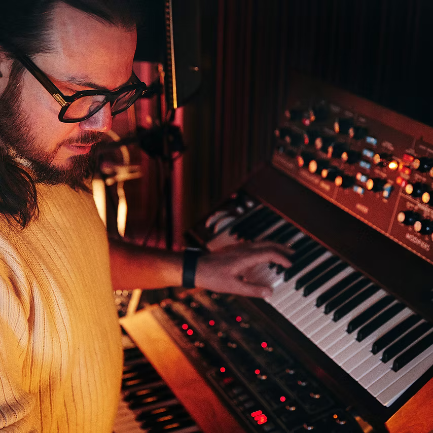 A man wearing glasses and a yellow shirt playing an electronic keyboard in a cozy music studio with warm lighting.