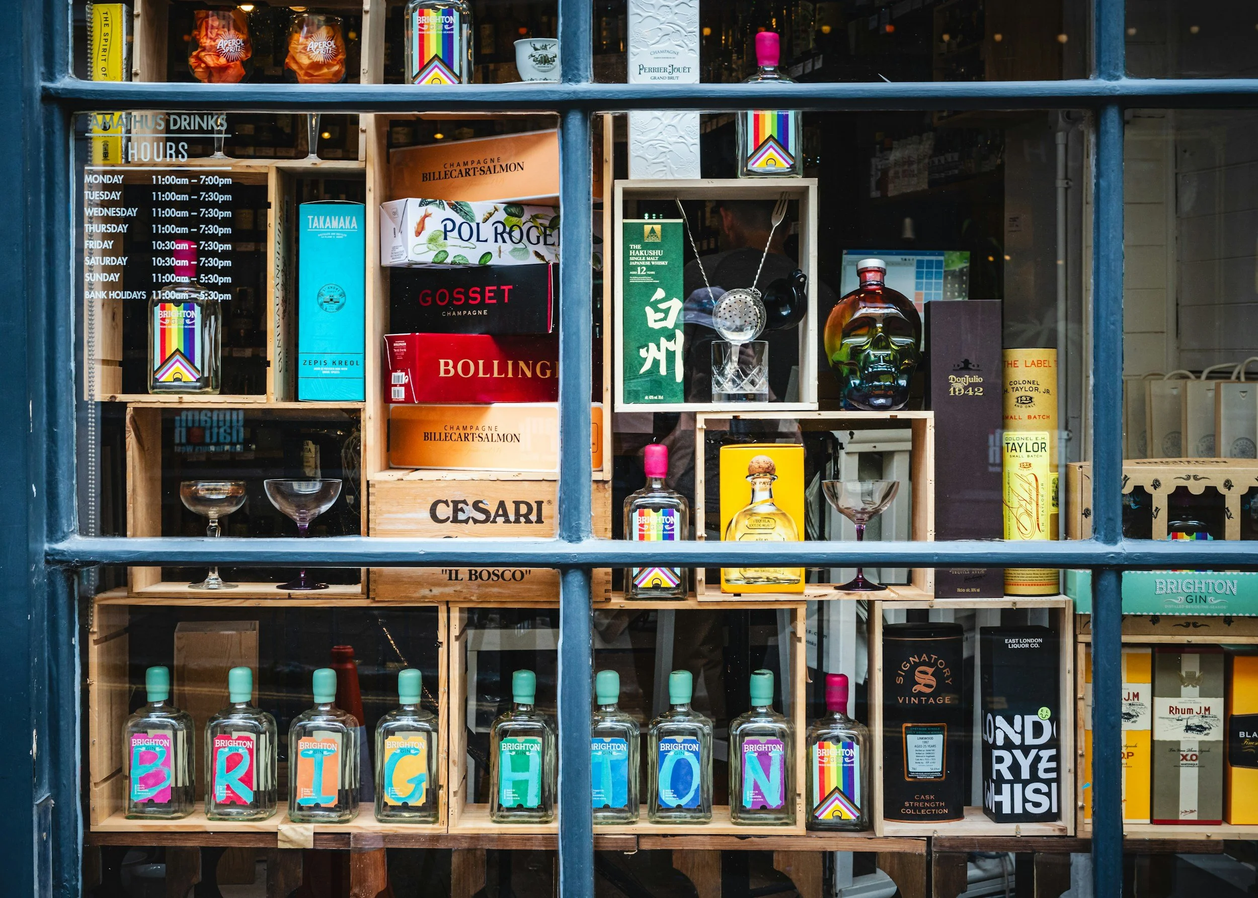 A store window display with wooden shelves holding bottles of Brighton gin, colorful boxes of liquor, and various decorative items including a rainbow-colored bottle, a skull-shaped bottle, and small glasses. The window also has printed hours of operation.