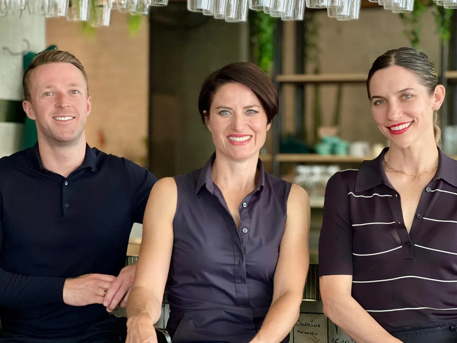 Three smiling people sitting at a table in a restaurant or cafe, with glasses hanging upside down from the ceiling above them.
