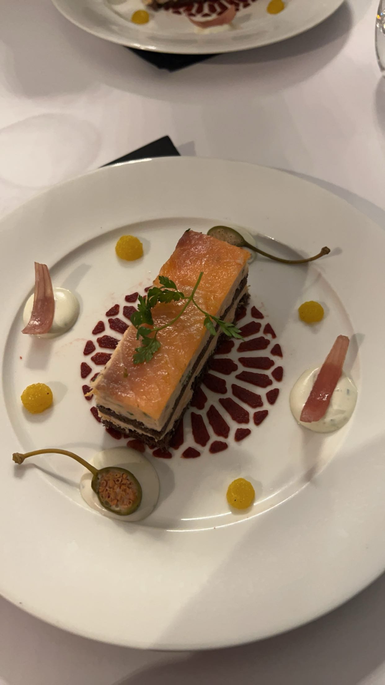 Assiette avec un dessert en forme de rectangle, décorée avec des petits pois jaunes, des gousses de figues, des morceaux de jambon cru, une sauce blanche et une sauce rouge avec des herbes.