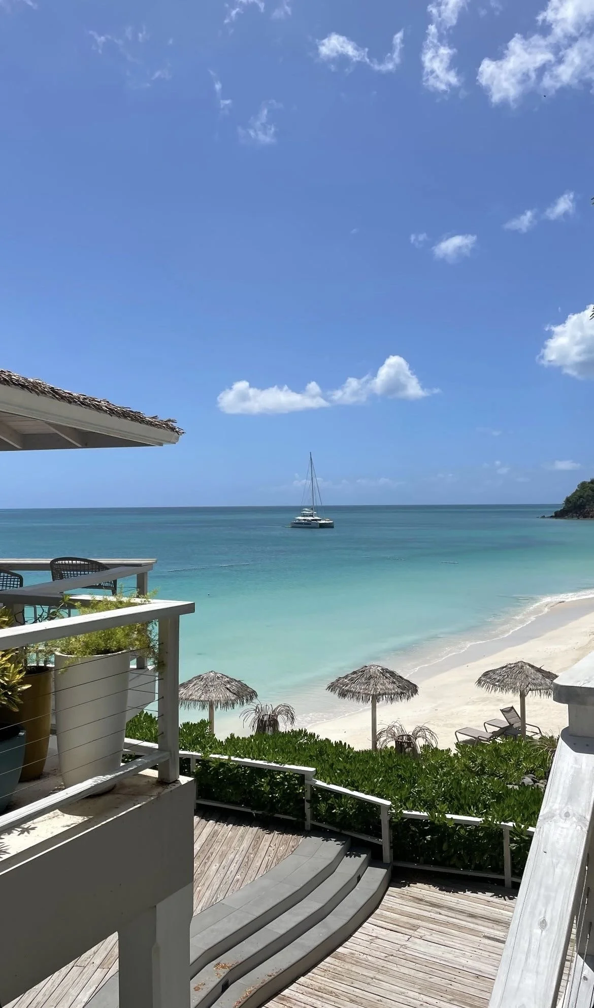 Plage de sable blanc avec parasols en paille, mer turquoise, ciel bleu avec quelques nuages, et un voilier au loin.