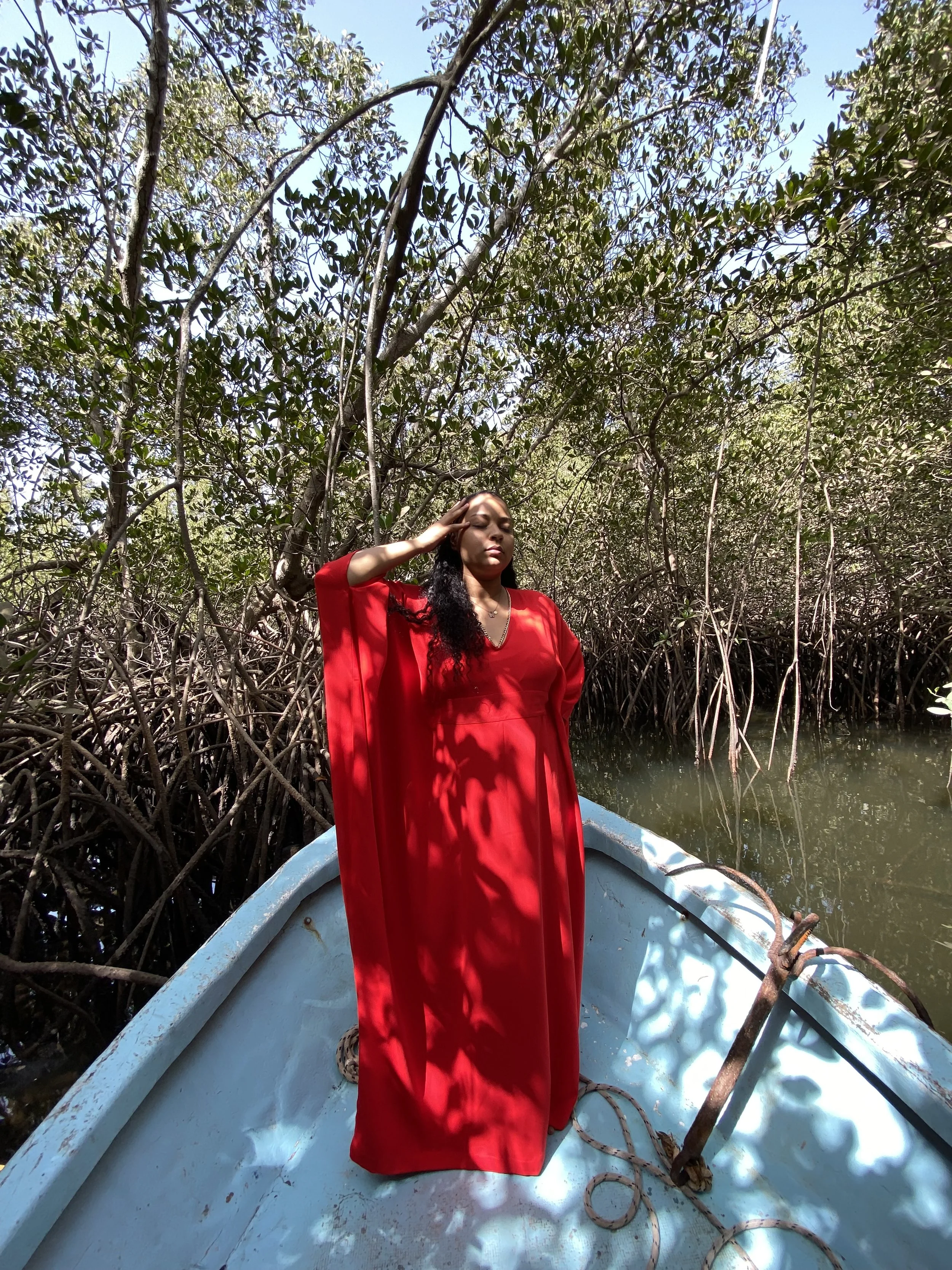 Femme en robe rouge debout sur un bateau dans une mangrove, saluant ou se protégeant du soleil, entourée de branches d'arbres et de l'eau.