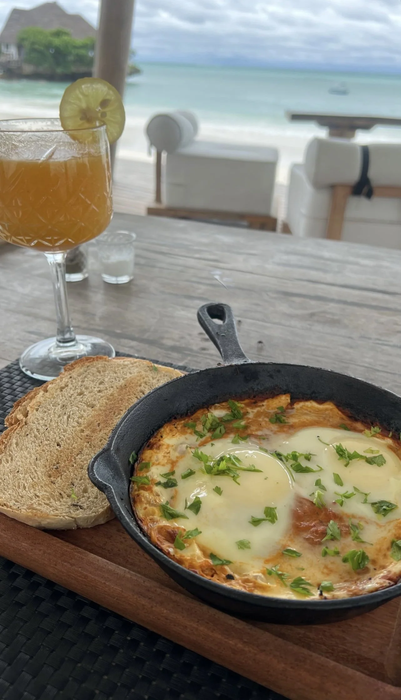 Oeuf au plat avec garniture verte, deux tranches de pain, verre de cocktail avec tranche de kiwi, décor de plage avec fauteuils et vue sur mer