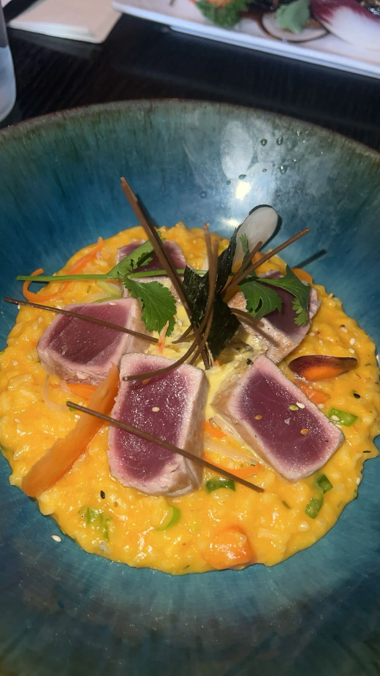 Un plat de tartare de thon cru servi sur un lit de risotto à la carotte, décoré avec des fines herbes, des légumes et des galettes de nori.