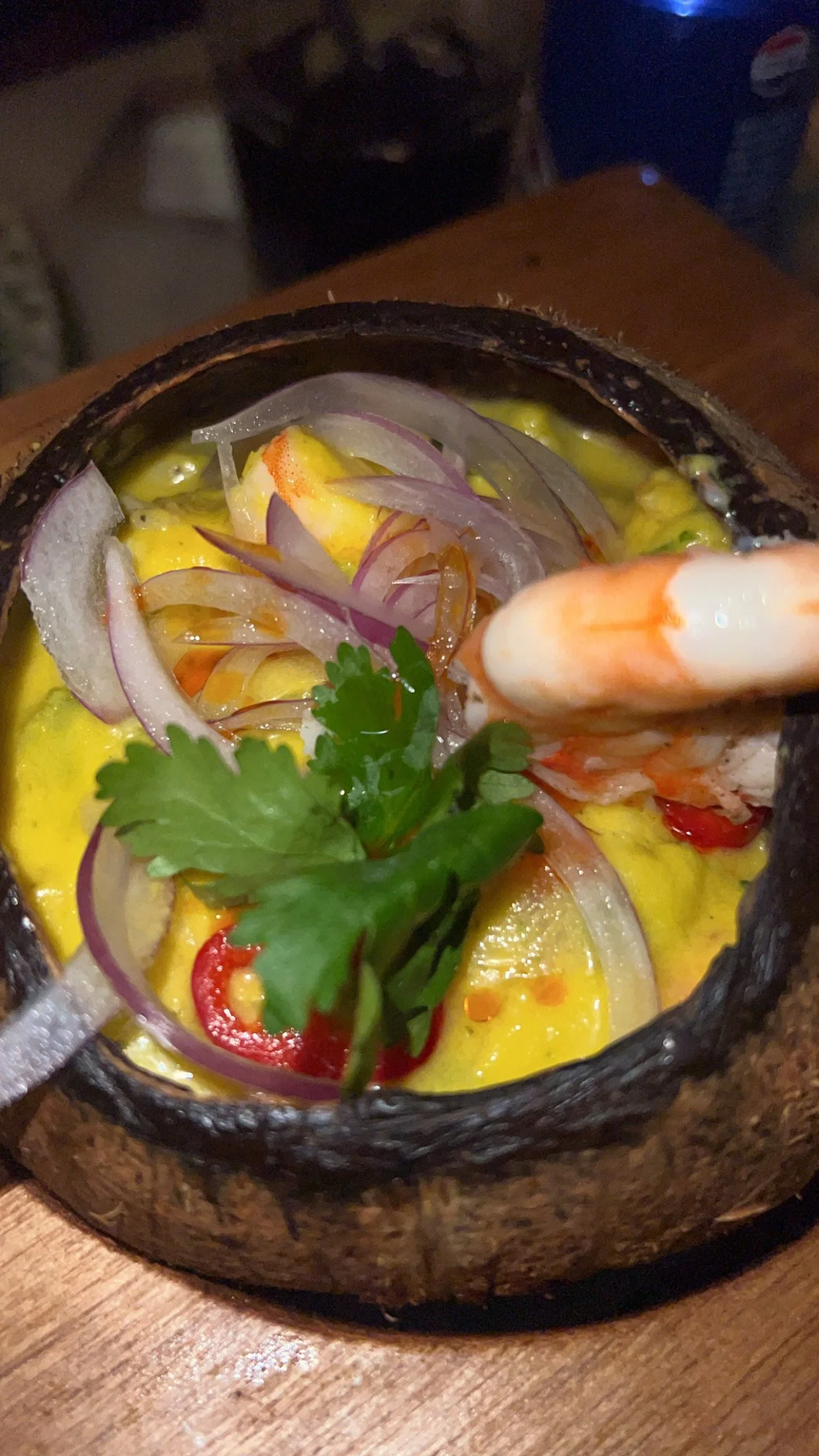 Soupe de crevettes servie dans un bol en pierre avec des oignons rouges, de la coriandre, des piments, du citron vert et une crevette visible.