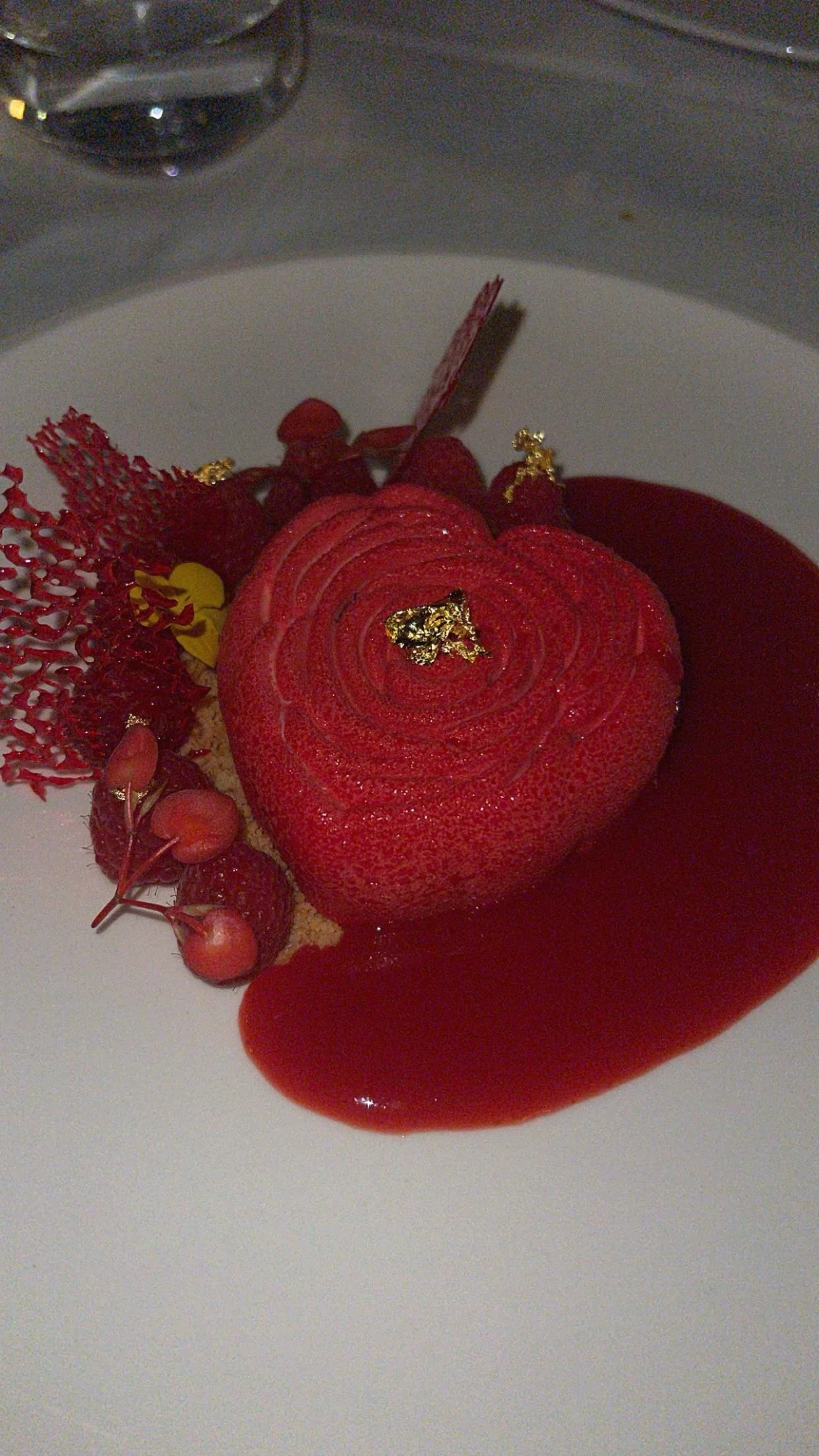Dessert en forme de cœur rouge, garni de décoration en or, avec de la compote de fruits rouges et des petits fruits rouges, présenté sur une assiette blanche.