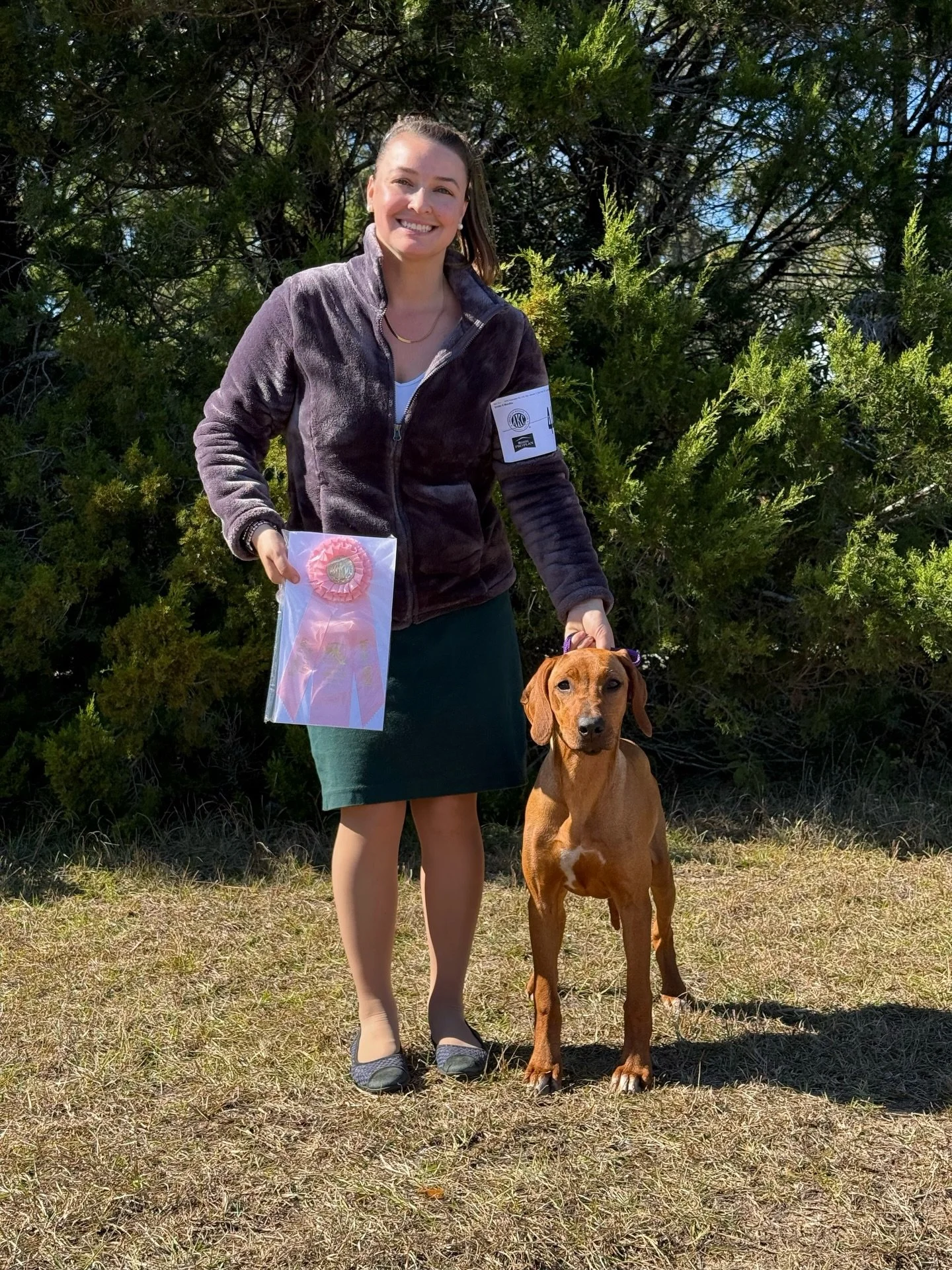 A young woman smiling and holding a pink ribbon rosette and a leash attached to a young Rhodesian ridgeback standing outdoors on grass in front of green bushes.