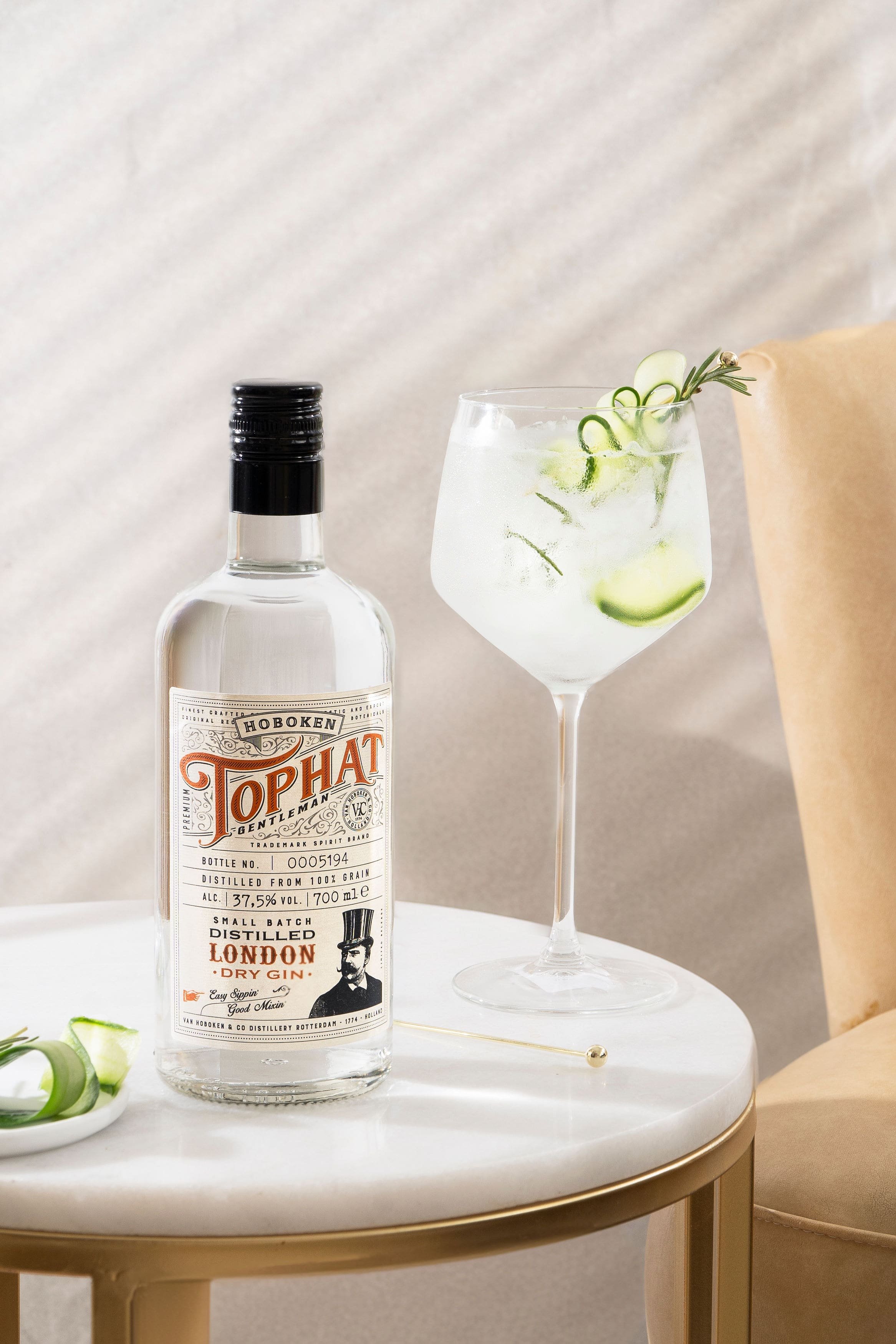 Bottle of Top Hat dry gin and a gin cocktail with cucumber garnish on a round table.