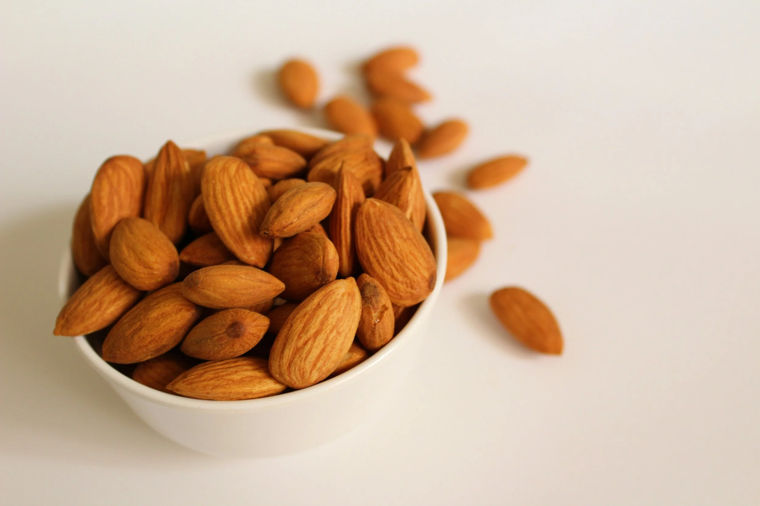 Whole almonds in a white bowl, with several almonds scattered nearby.