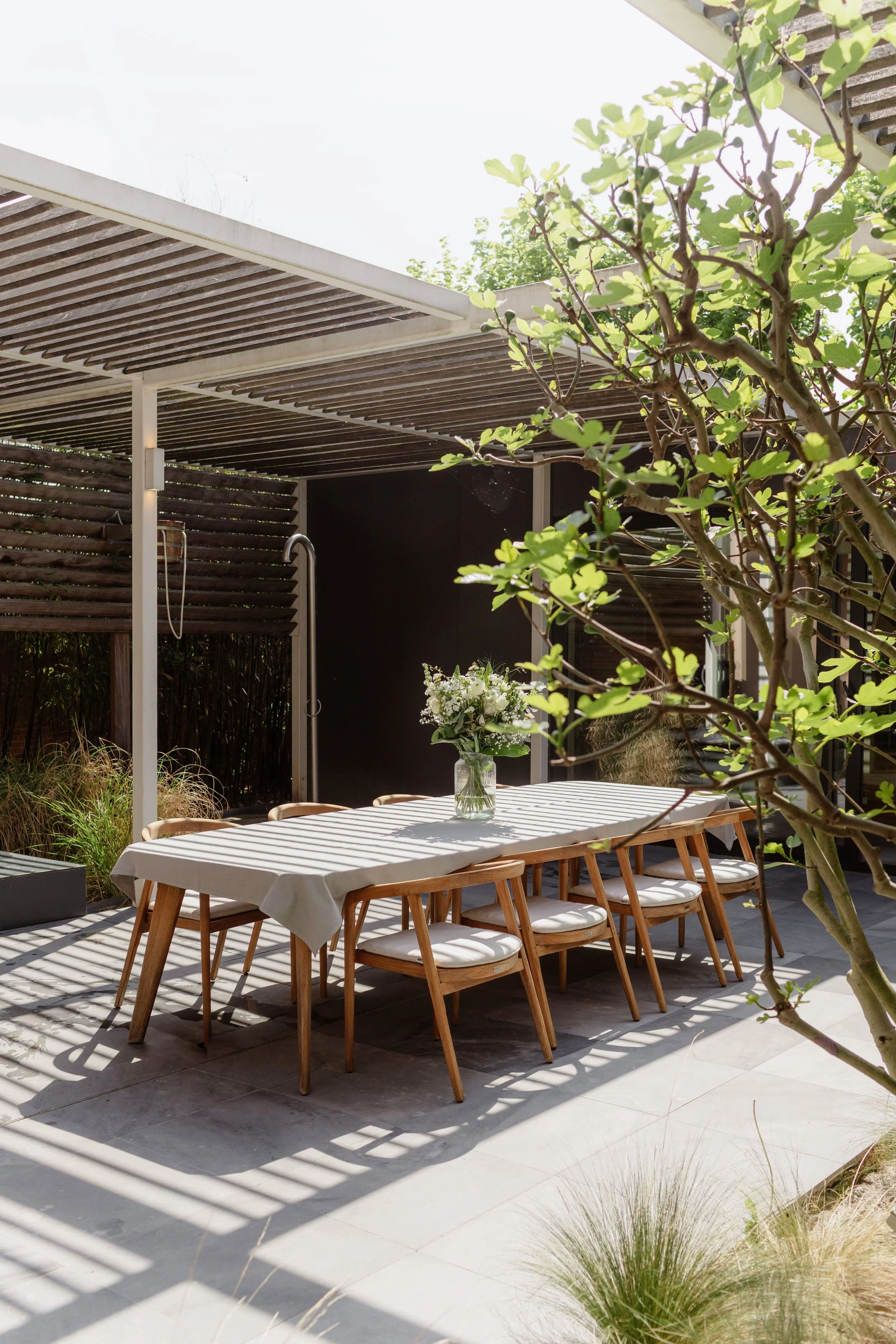 Eetstafel met tafelkleed en bloemstuk in een moderne tuin met schaduw op de terrasvloer.