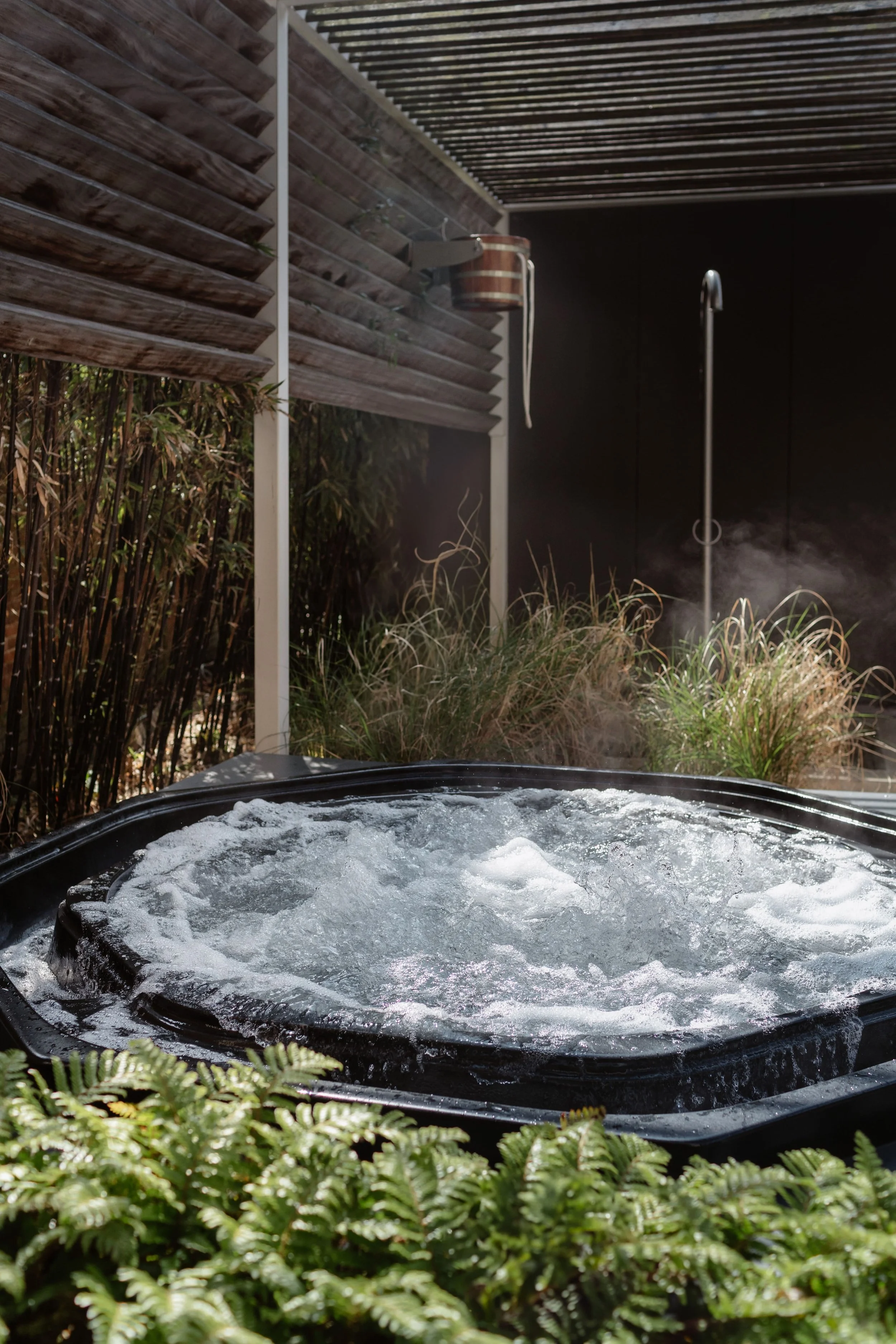 Buiten bubbelbad met stoom, omgeven door groene planten en houten schutting, met een douchekop op achtergrond.