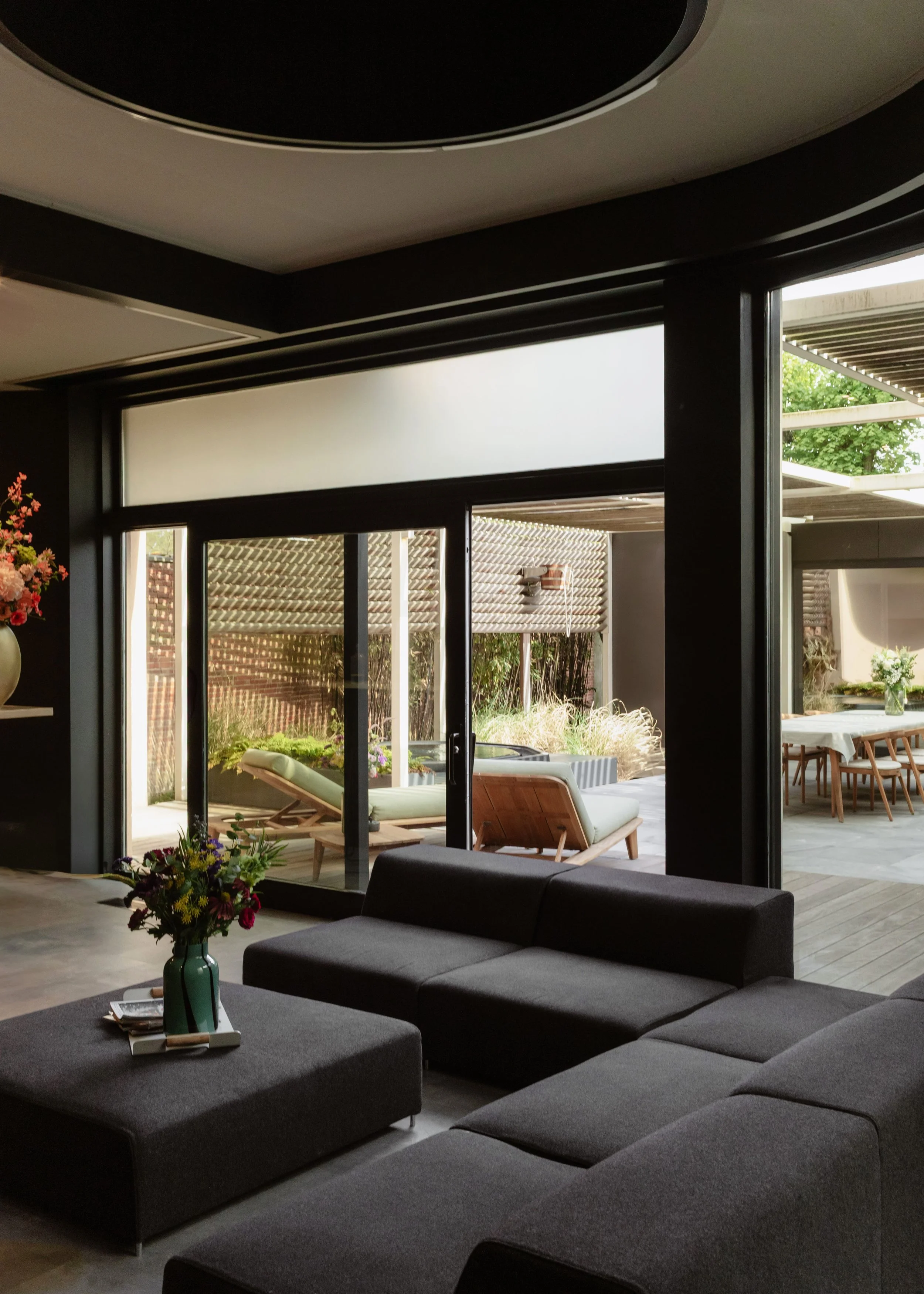 Uitzicht op een woonkamer met grote glazen schuifdeuren die leiden naar een tuin, met een zwarte bank en een bloemenvaas op een salontafel.