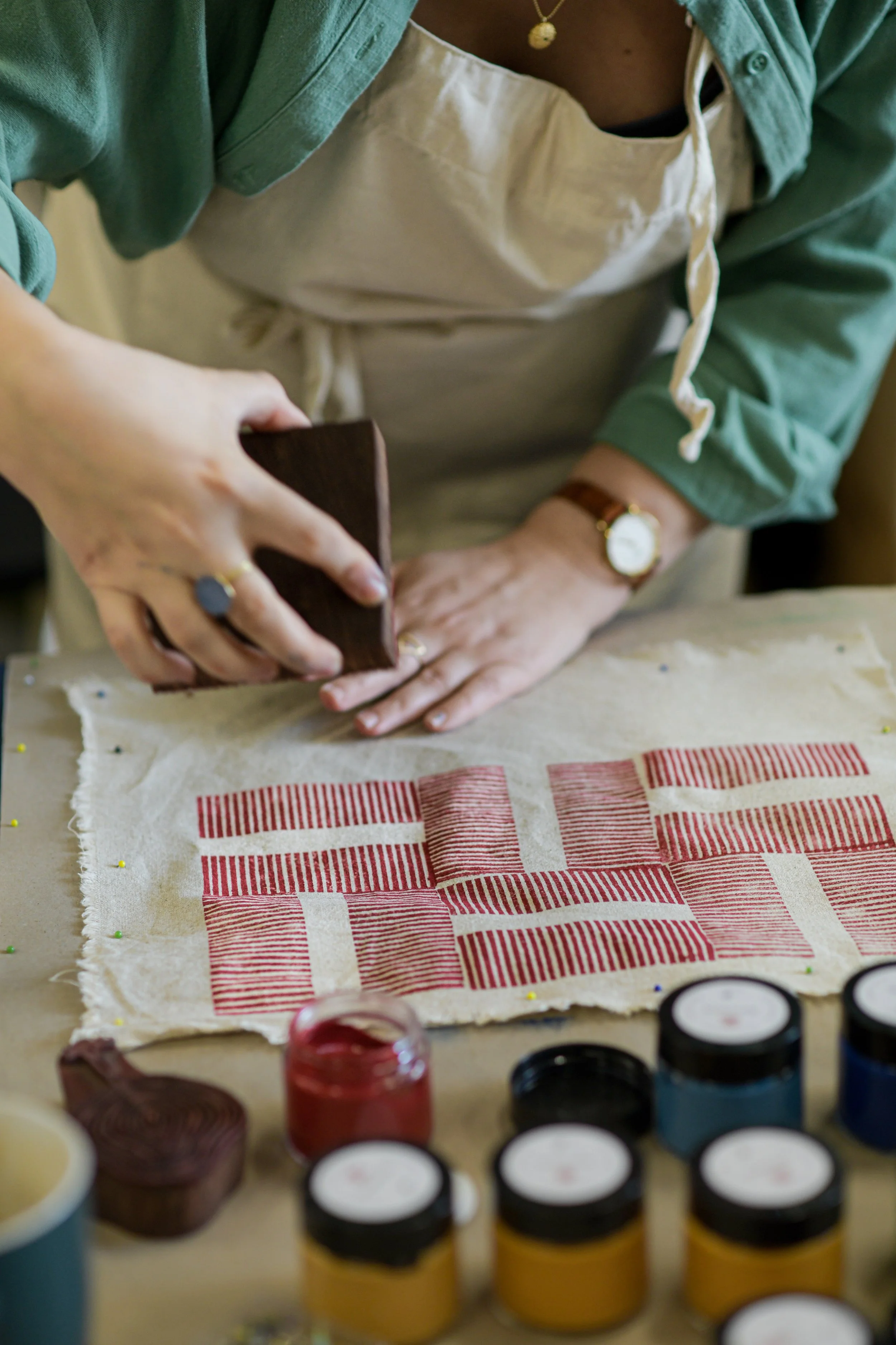 A person wearing a green shirt, beige apron, and a watch, working on a block printing project on a table at Atelier Māya in Berlin. Several jars of fabric dye and containers are nearby.