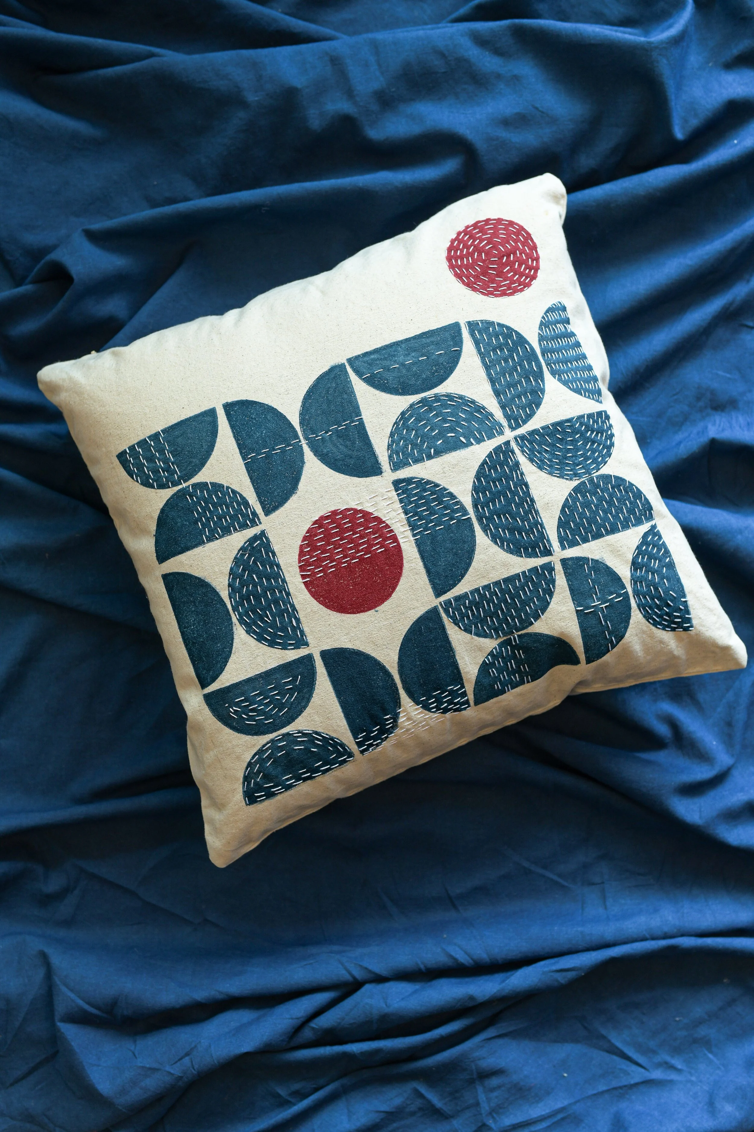 A decorative pillow with abstract navy blue shapes and red accents, resting on a crinkled dark blue fabric background.
