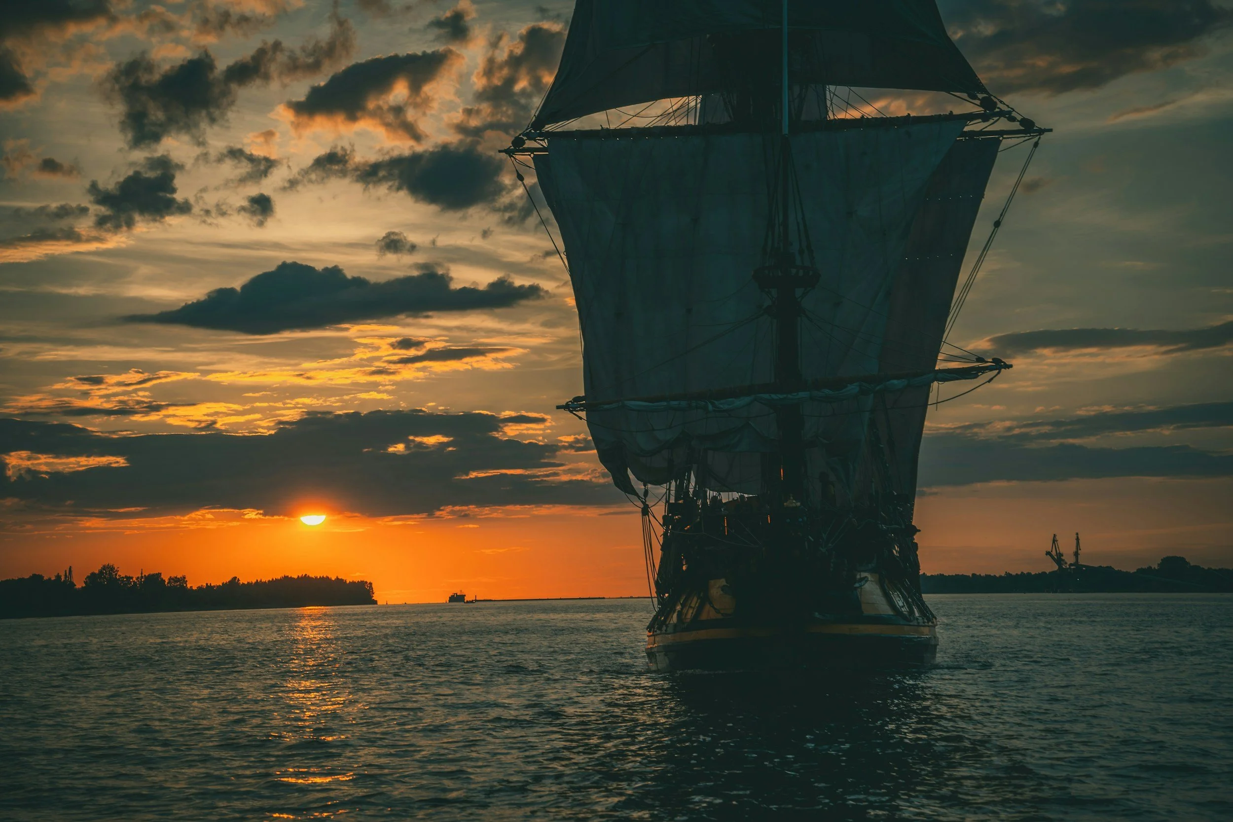 A sailing ship with raised sails on the water during sunset, with a partly cloudy sky and a silhouette of land and cranes in the background.